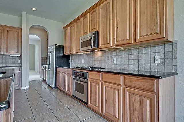 Kitchen featuring wood cabinetry, dark countertops, tiled backsplash, stainless steel appliances, and tiled flooring