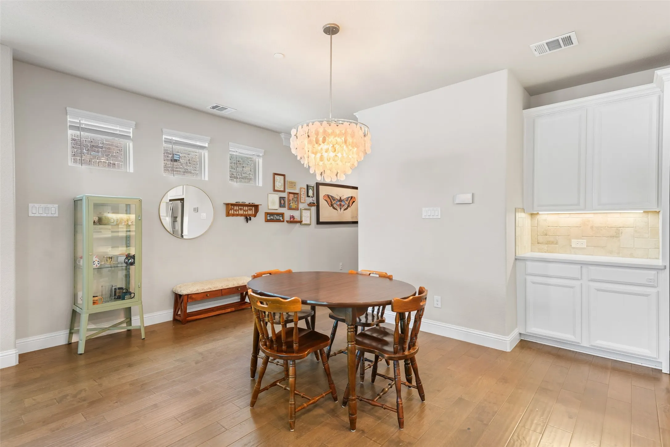 Dining area featuring wood-finish flooring, a contemporary shell chandelier, built-in cabinetry with under-cabinet lighting, a light-toned tile backsplash, and multiple high-set windows