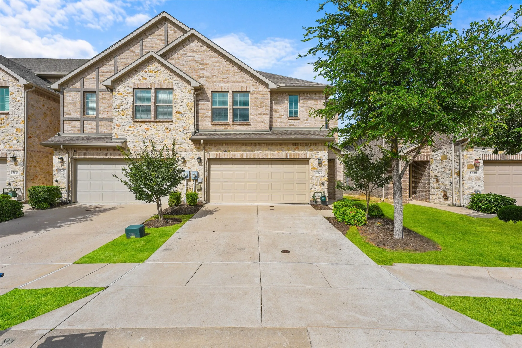 Two-story stone and brick facade townhome featuring an attached two-car garage, concrete driveway, and established landscaping