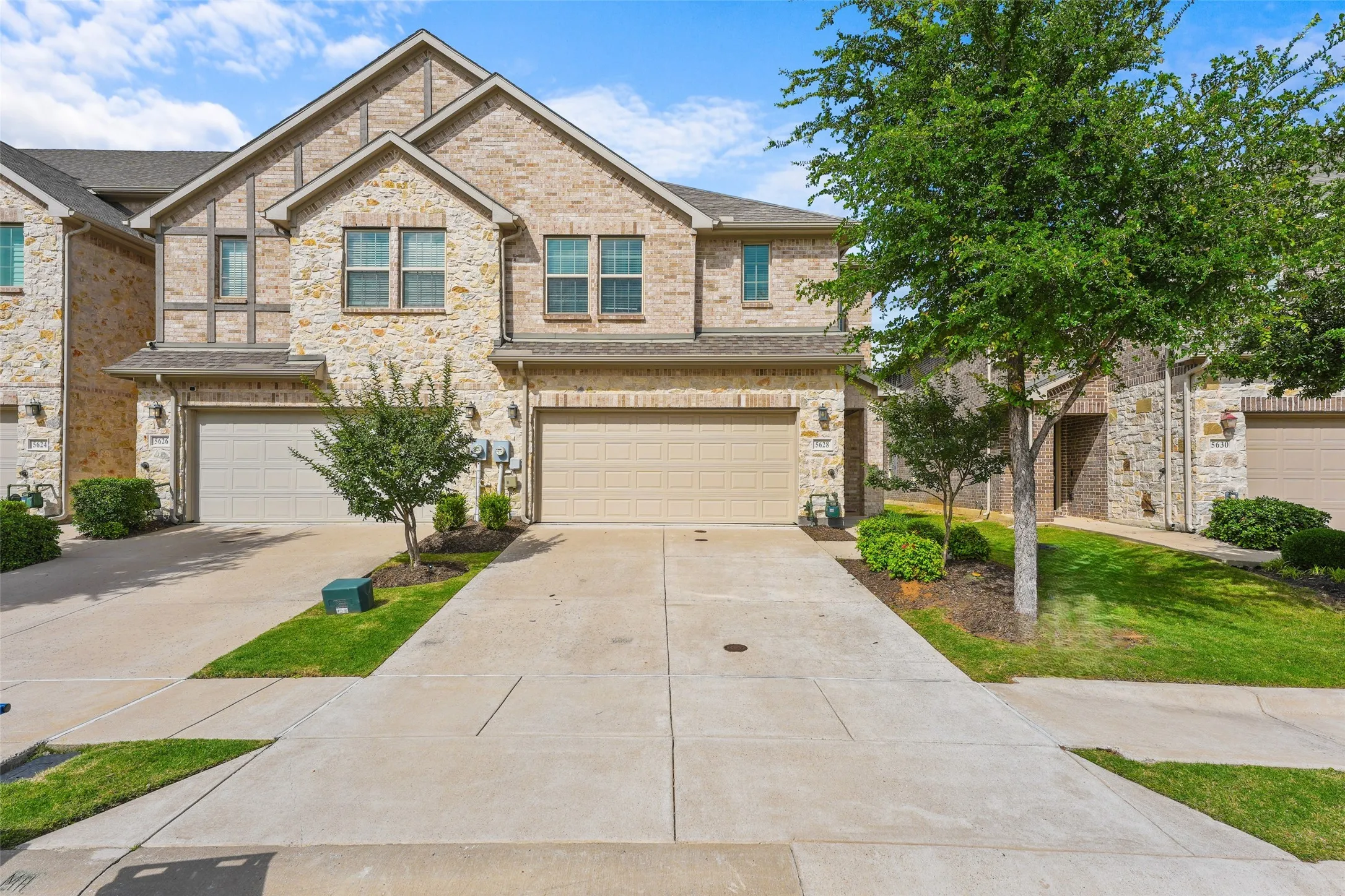 Stone facade townhome featuring an attached garage, paved driveway, mature trees, and landscaped shrubbery