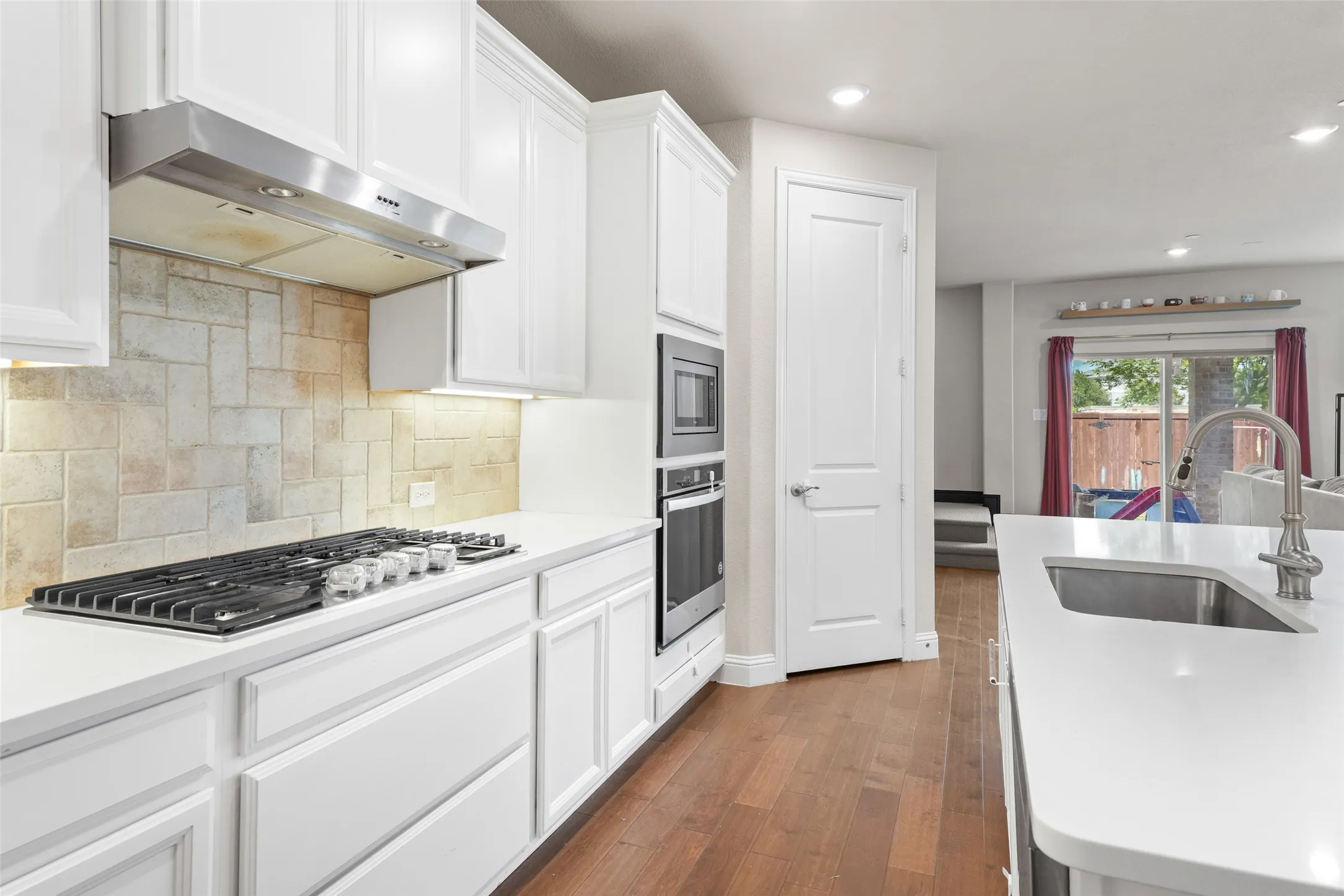 Bright kitchen featuring white cabinetry, stone-look backsplash, stainless steel range hood, built-in oven and microwave, and a kitchen island with an undermount sink and gooseneck faucet