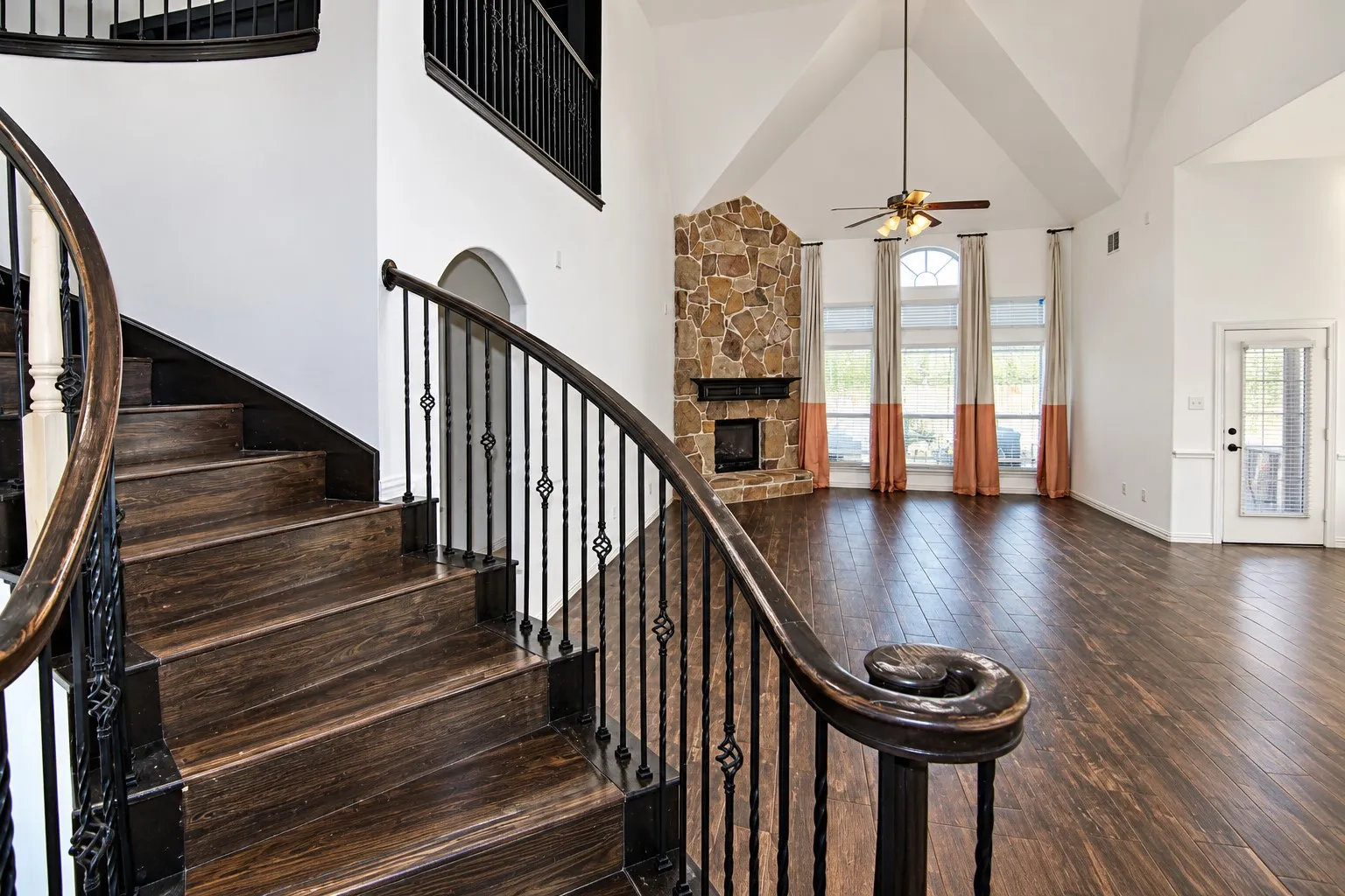Vaulted ceiling, stone-clad fireplace, wood-finish flooring, curved staircase with wrought iron balusters, and arched windows