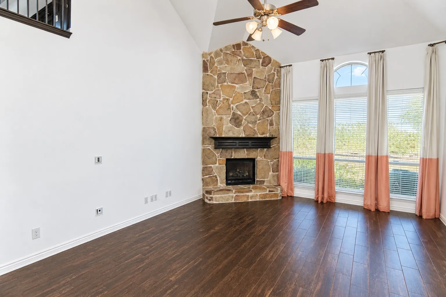 Vaulted ceiling with a ceiling fan, stone-clad fireplace, wood-finish flooring, and multiple tall windows with an arched transom