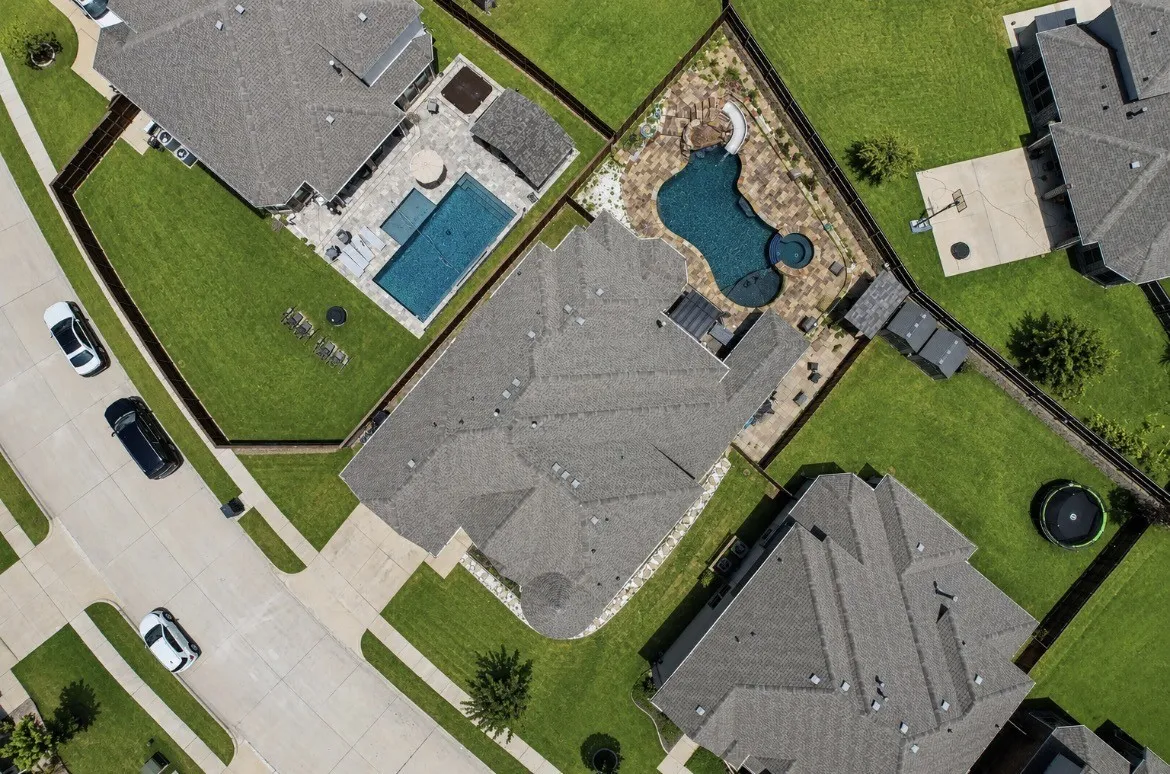 Aerially captured property showcasing a curvilinear swimming pool with integrated spa, a paved patio surround, and an adjacent covered outdoor structure