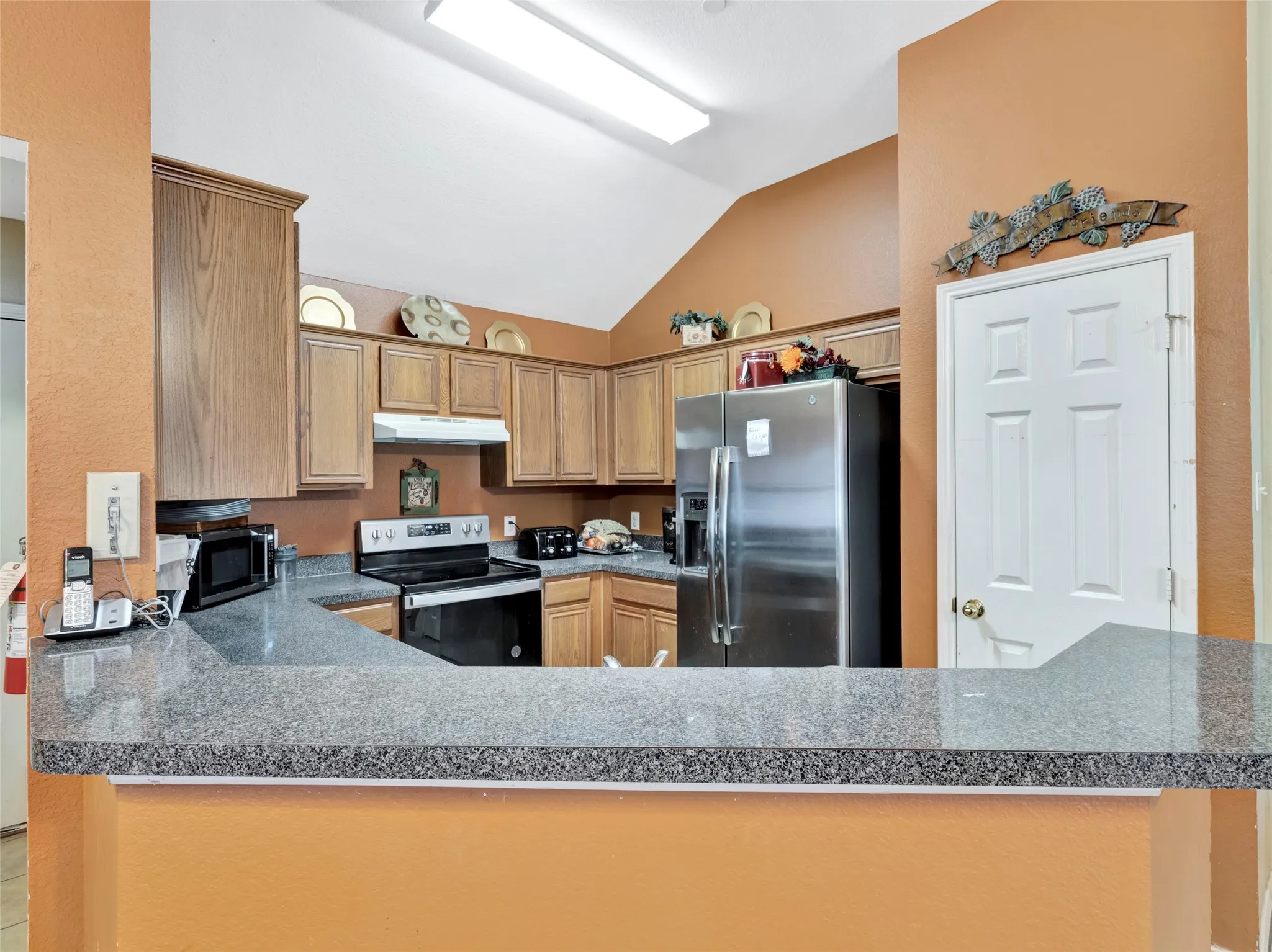 Vaulted ceiling, wood-finish cabinetry, granite-finish countertops, stainless steel refrigerator, and built-in electric range