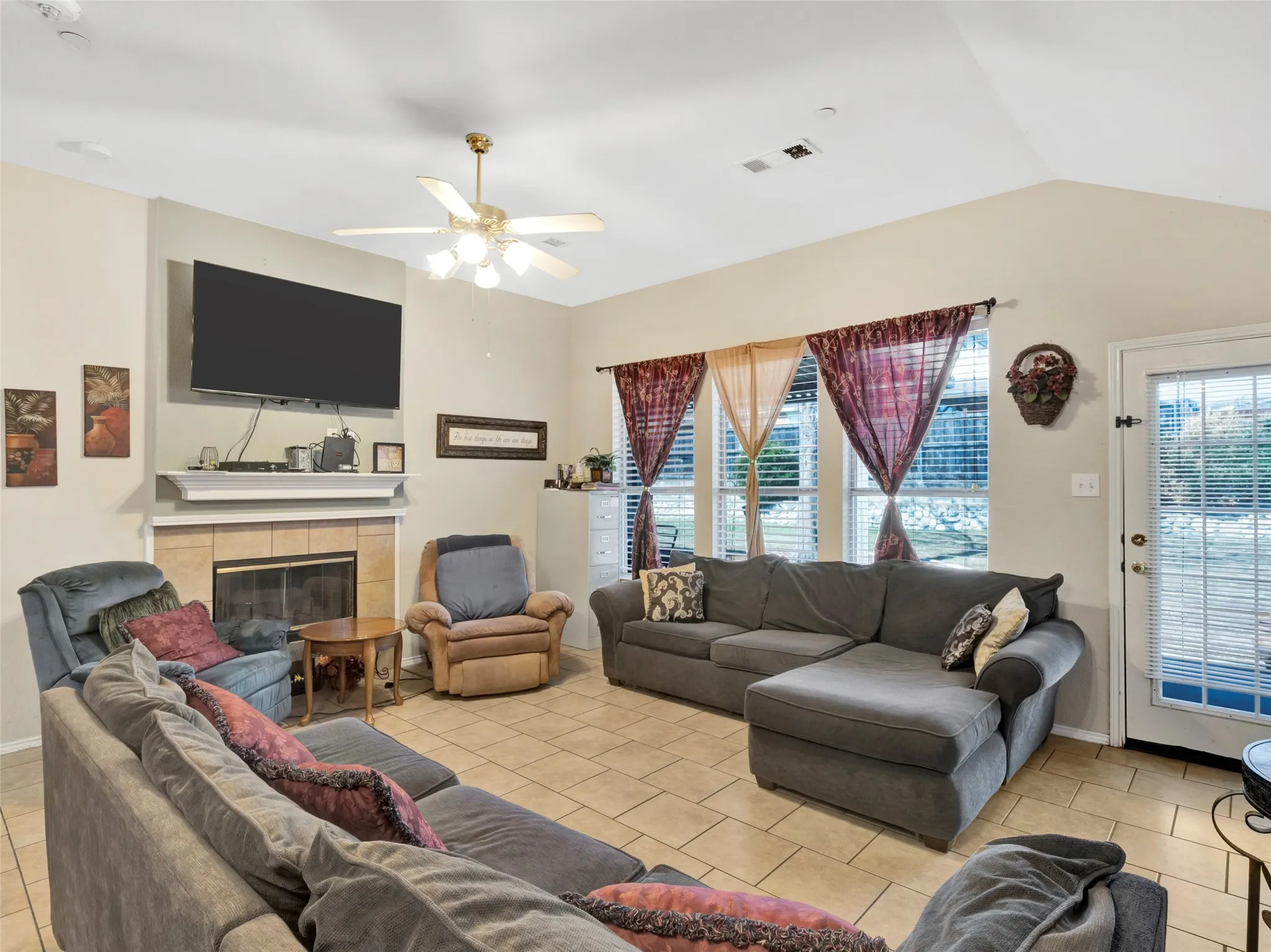 Vaulted ceiling, ceiling fan with light fixture, tiled fireplace with mantel, tiled flooring, and a glass-paneled door