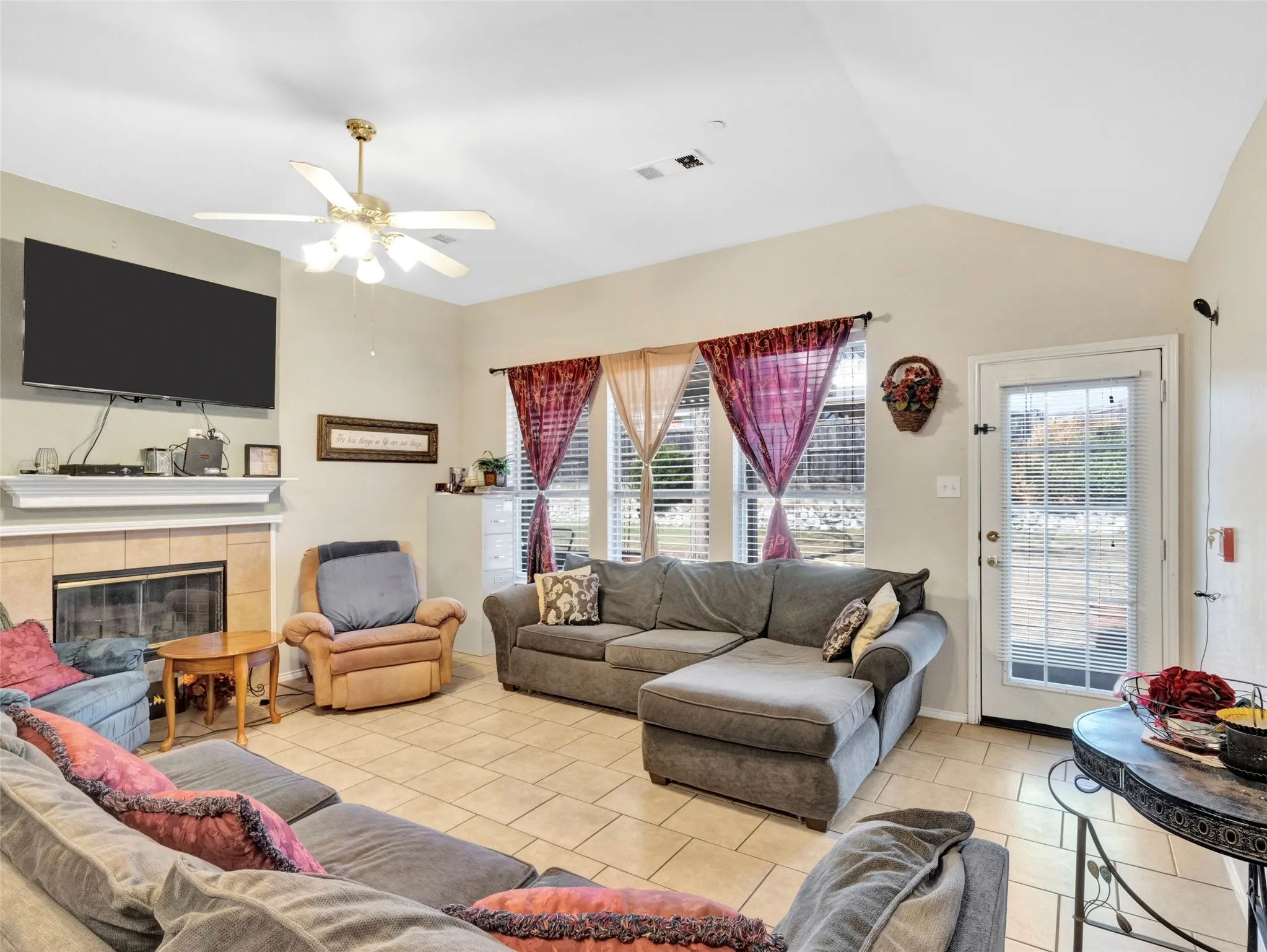 Vaulted ceiling with a ceiling fan, tiled flooring, fireplace with a tiled surround and mantel, multiple windows with blinds, and a glass-paneled door
