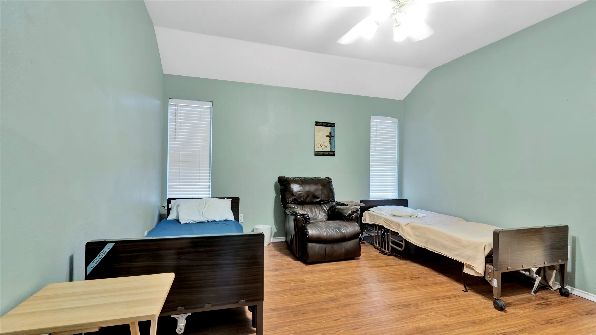 Vaulted ceiling with a ceiling fan, light green wall paint, wood-finish flooring, and two windows with blinds