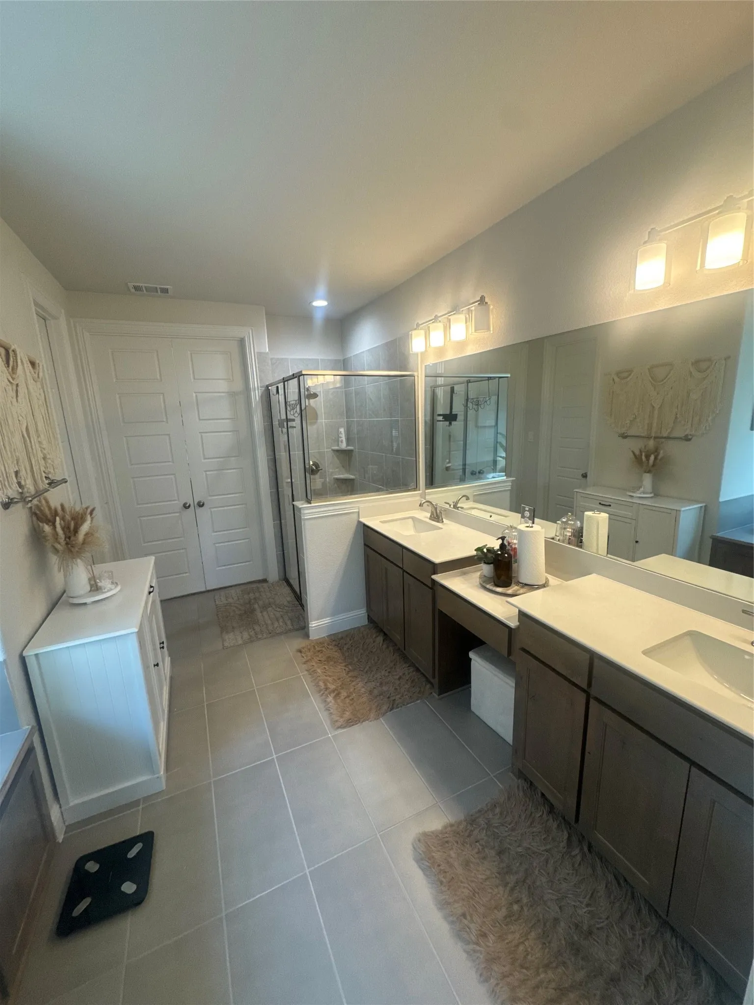 Bathroom featuring dual vanities with integrated sinks, a built-in shower with glass enclosure, light-toned floor tile, and double panel doors