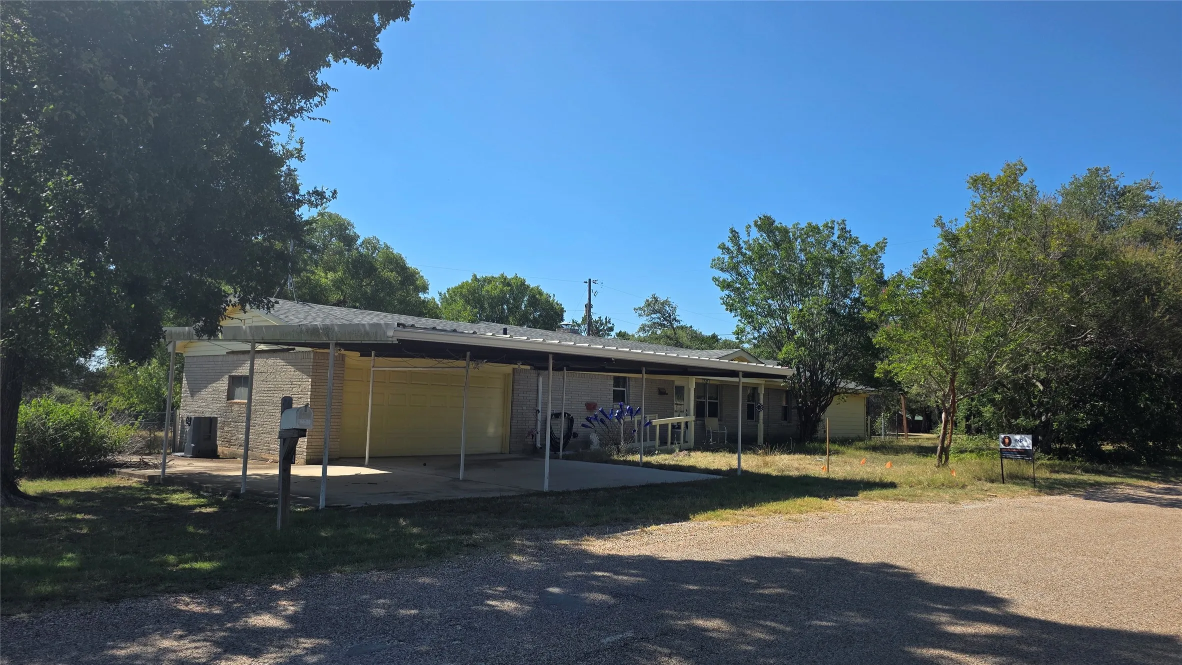 Single-story residence featuring light-colored brick siding, a covered carport, an extended covered porch, a gravel driveway, and mature trees