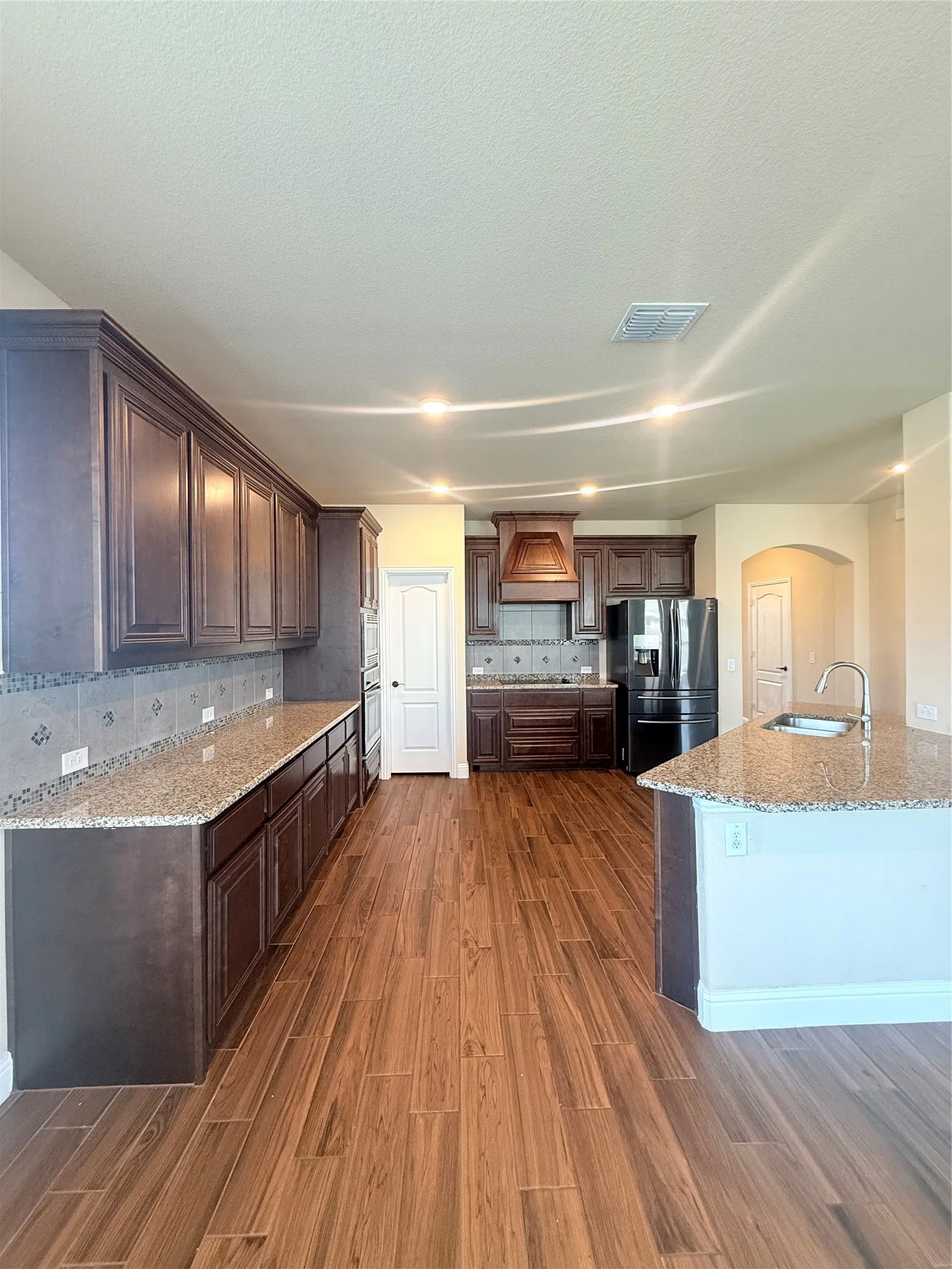 Spacious kitchen featuring rich wood cabinetry, granite countertops, tile backsplash, a large central island with sink, and wood-finish flooring