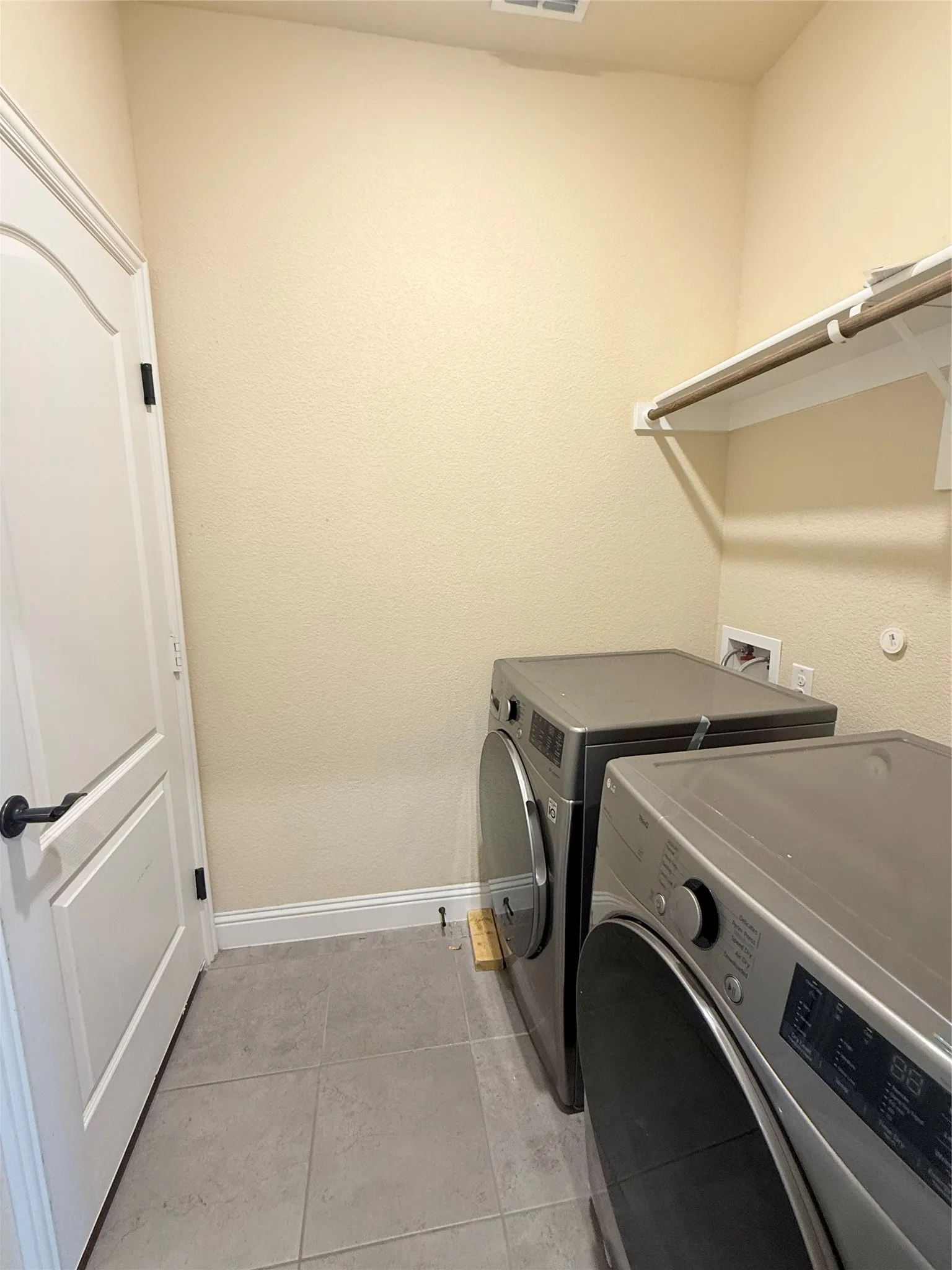 Utility area featuring light beige wall finish, grey tiled flooring, white baseboards, and a wall-mounted shelving unit