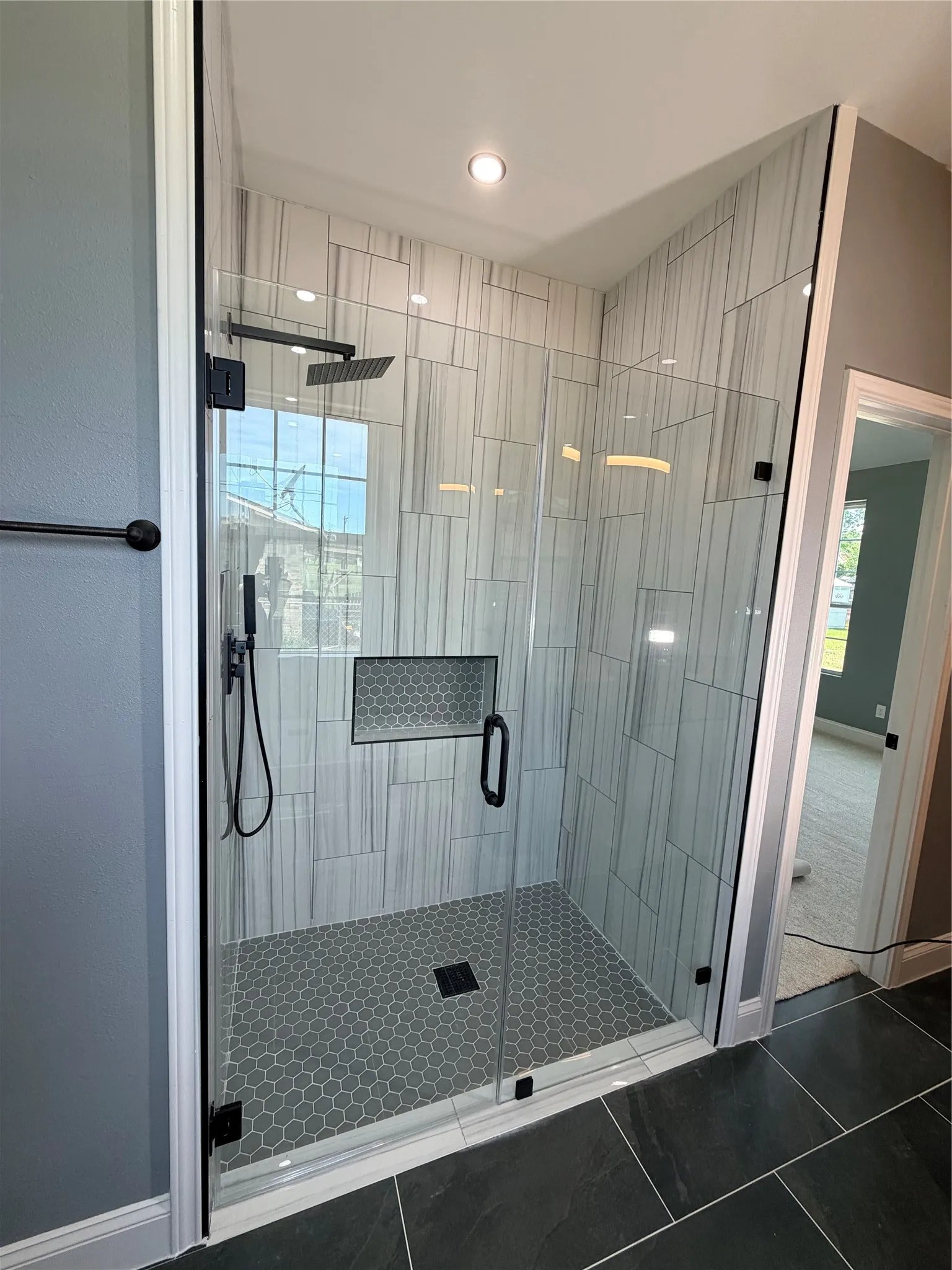 Frameless glass shower enclosure featuring a matte black rainfall showerhead, handheld sprayer, geometric mosaic tile shower niche, and hexagonal floor tiles