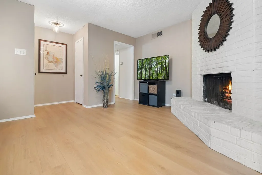 Expansive living area featuring light wood-finish flooring, a white-painted brick fireplace with a hearth, and neutral wall tones