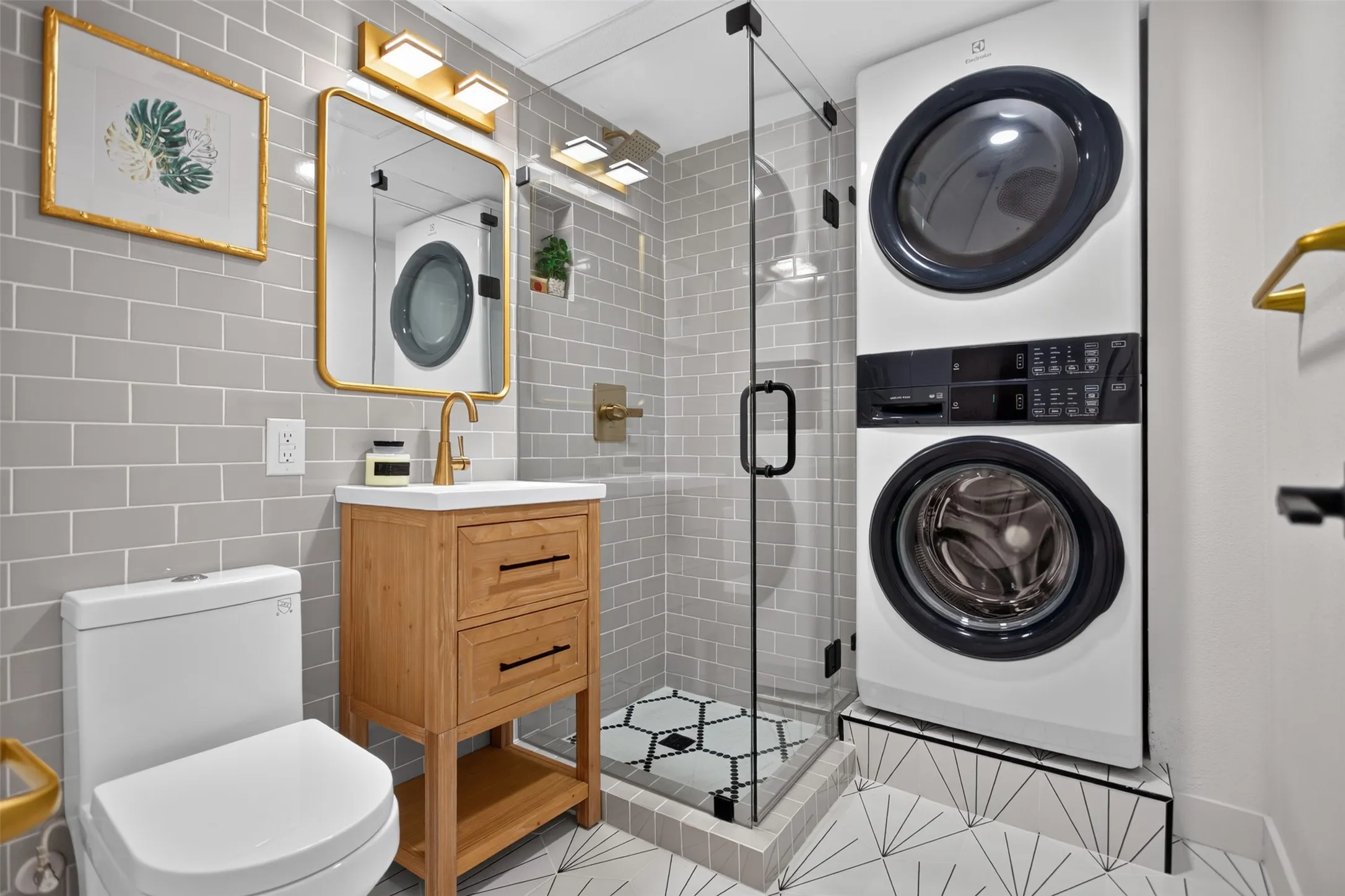 Bathroom featuring a  dryer unit, seamless glass shower with geometric floor tile, light gray subway tile from floor to ceil and wall to wall, a wood-finish vanity with a white countertop, and gold-finish fixtures