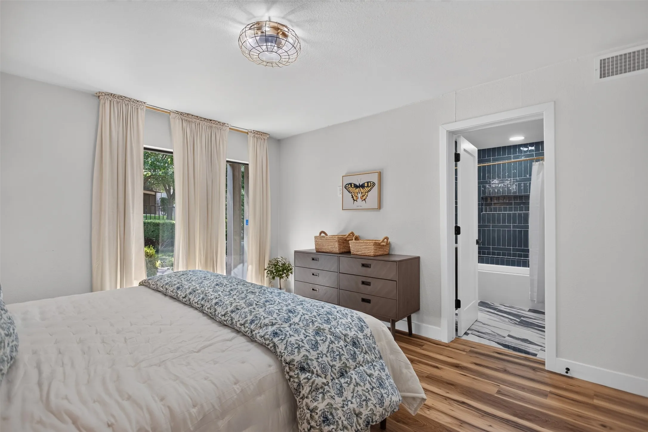 Bedroom featuring wood-finish flooring, two large windows with curtain rods, a ceiling-mounted light fixture, and an attached full bathroom with a tiled shower/tub combination