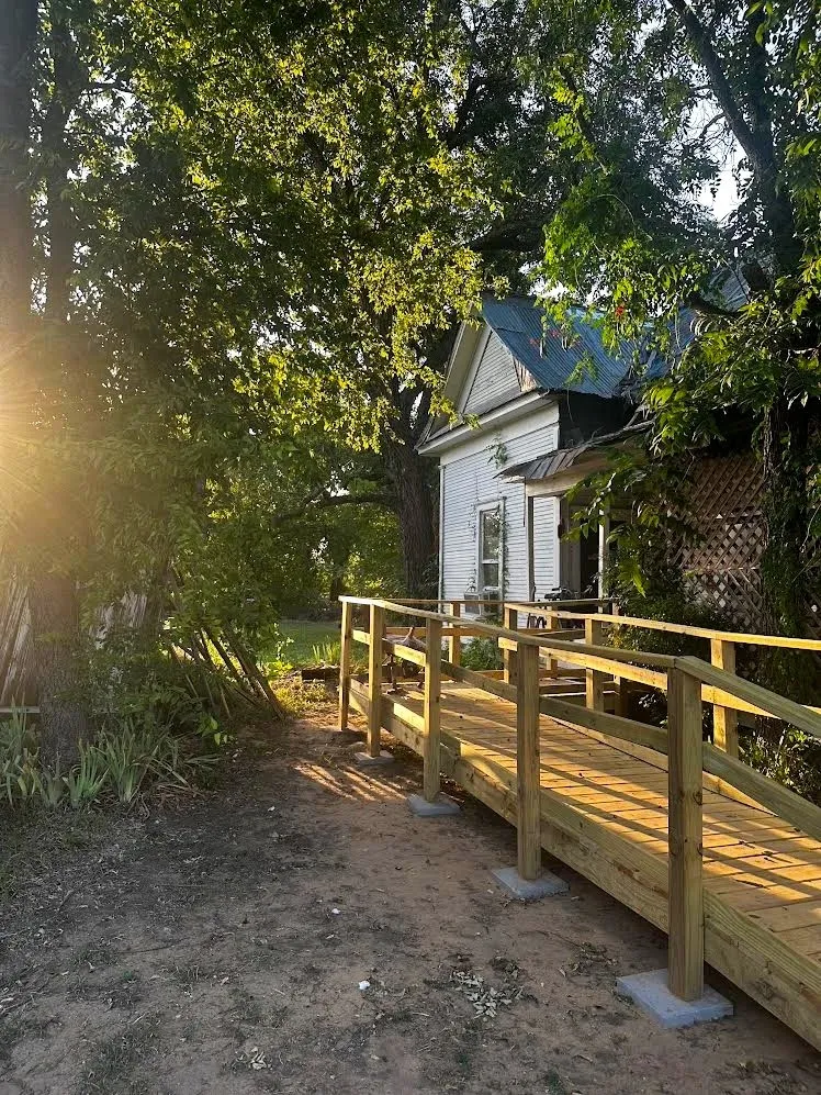 Exterior featuring a white siding finish, a prominent wooden accessibility ramp with handrails, and mature trees providing natural shade