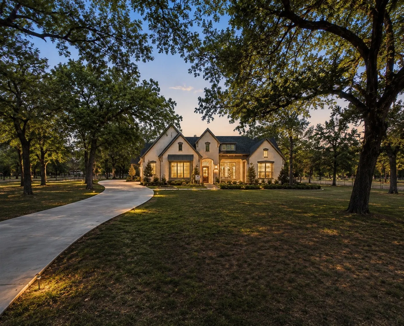 Elegant two-story residence featuring a light-colored stone and stucco exterior with arched entryway, multiple gabled rooflines, and substantial front-facing windows with warm interior lighting