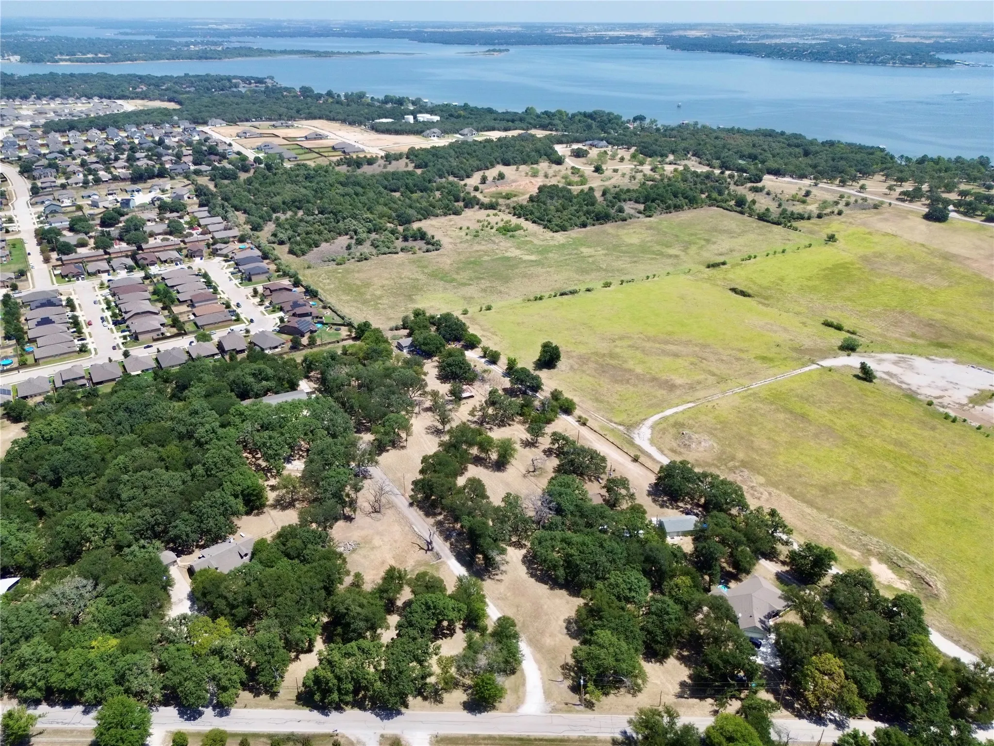 Expansive aerial view showcasing a large body of water, a residential neighborhood with numerous homes, significant green spaces with mature trees, open fields, and unpaved paths