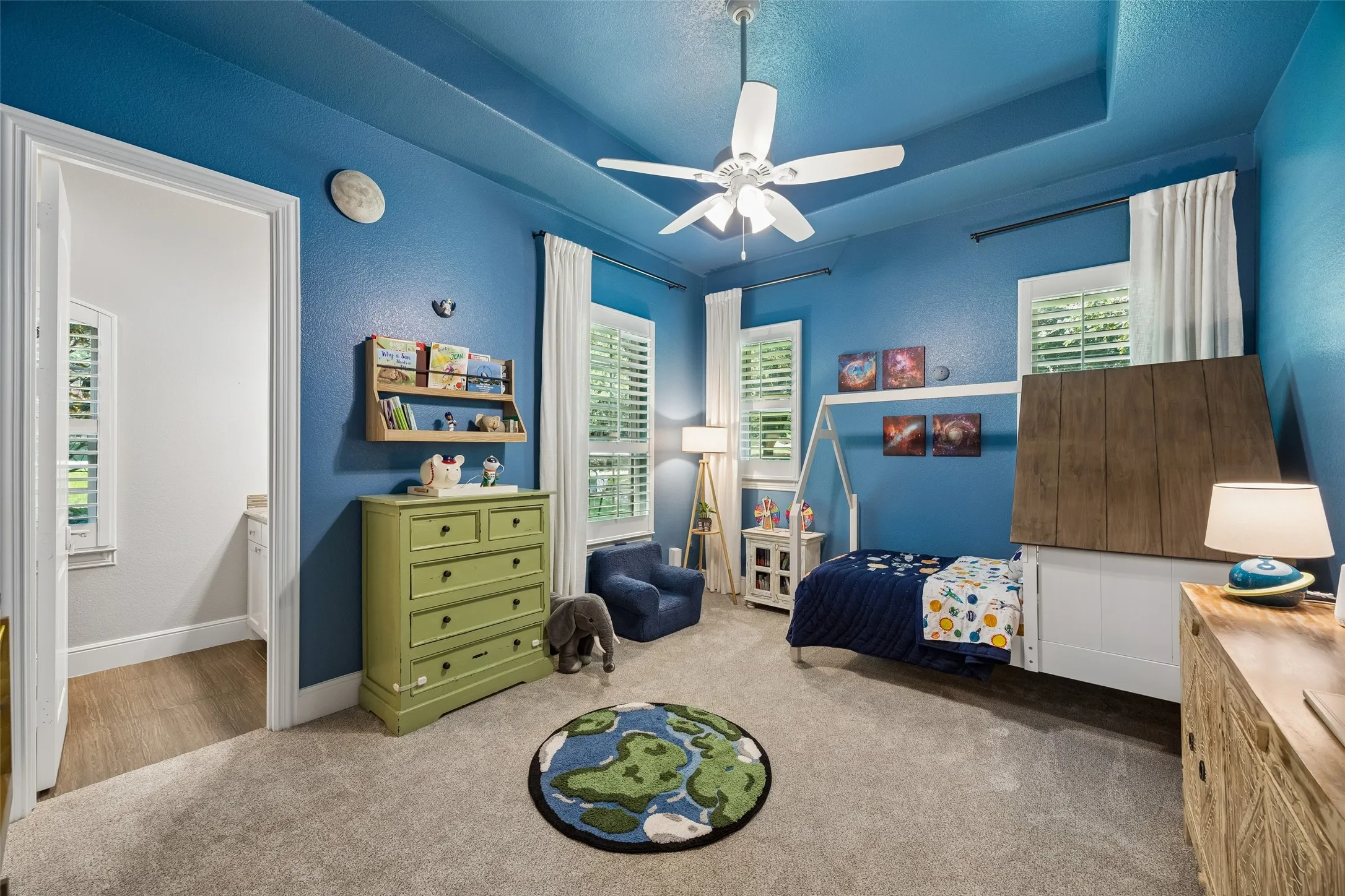 Carpeted room featuring a tray ceiling with a ceiling fan, two windows with plantation shutters, and a doorway leading to a separate area with wood-finish flooring