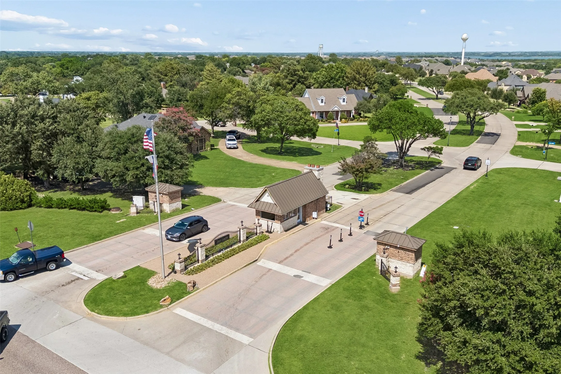 Gated community entrance featuring a stone and wrought iron entry monument, a guardhouse with a shingled roof, and a meticulously maintained landscape with mature trees