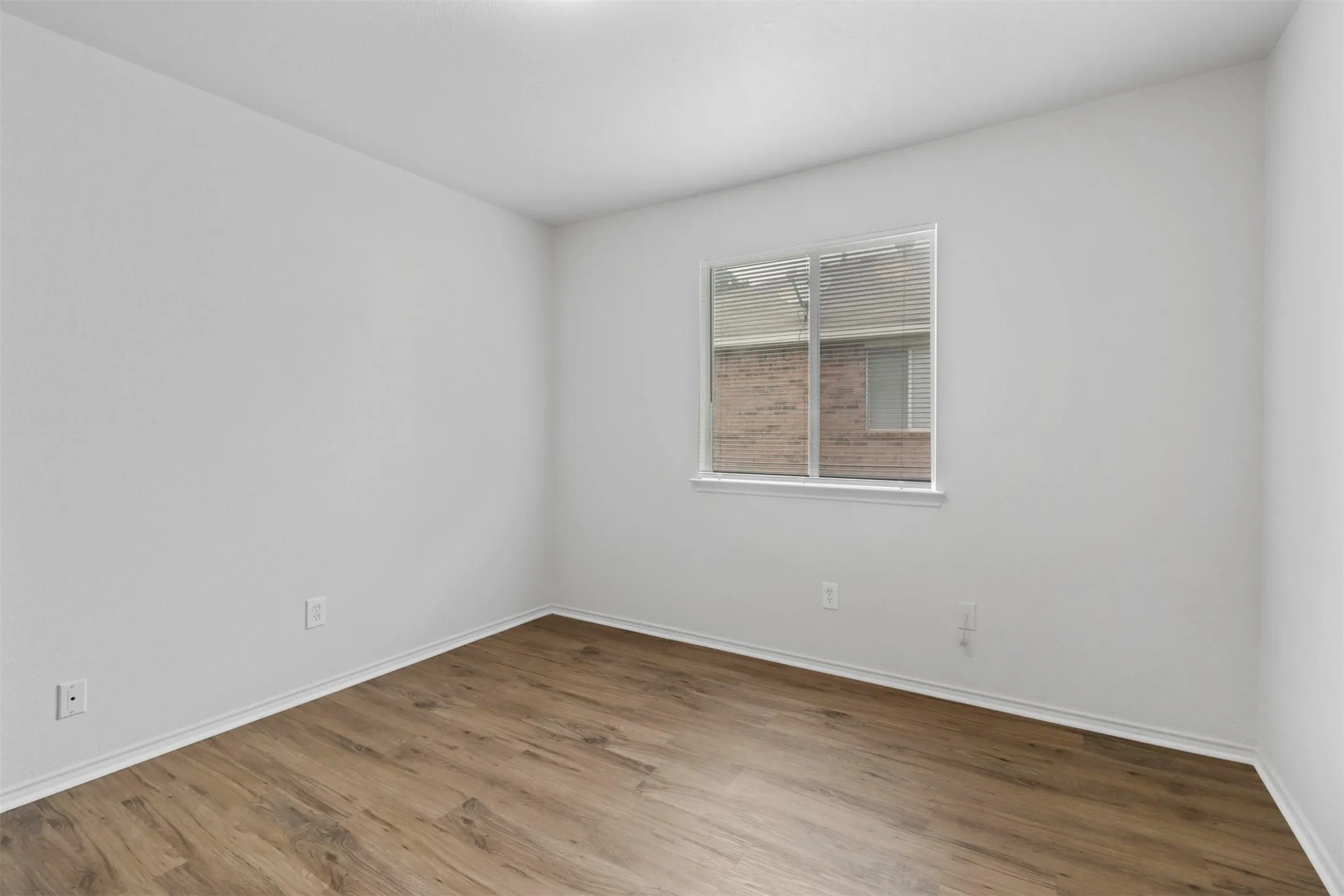 Room with light wood-finish flooring, white walls, white baseboards, and a single window featuring horizontal blinds