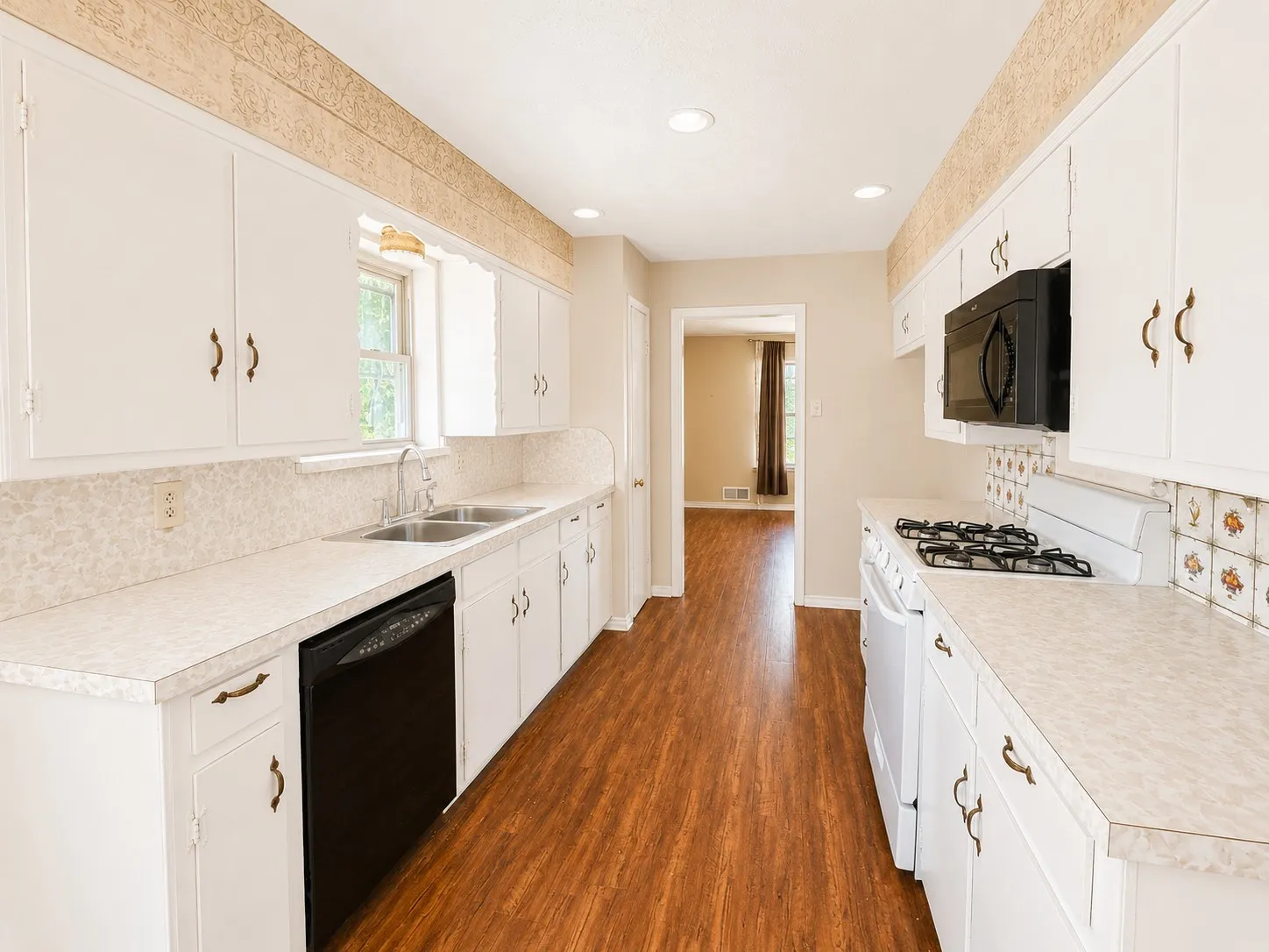 Galley kitchen featuring wood-finish flooring, white cabinetry with brass-tone hardware, a double basin sink, and a black built-in microwave