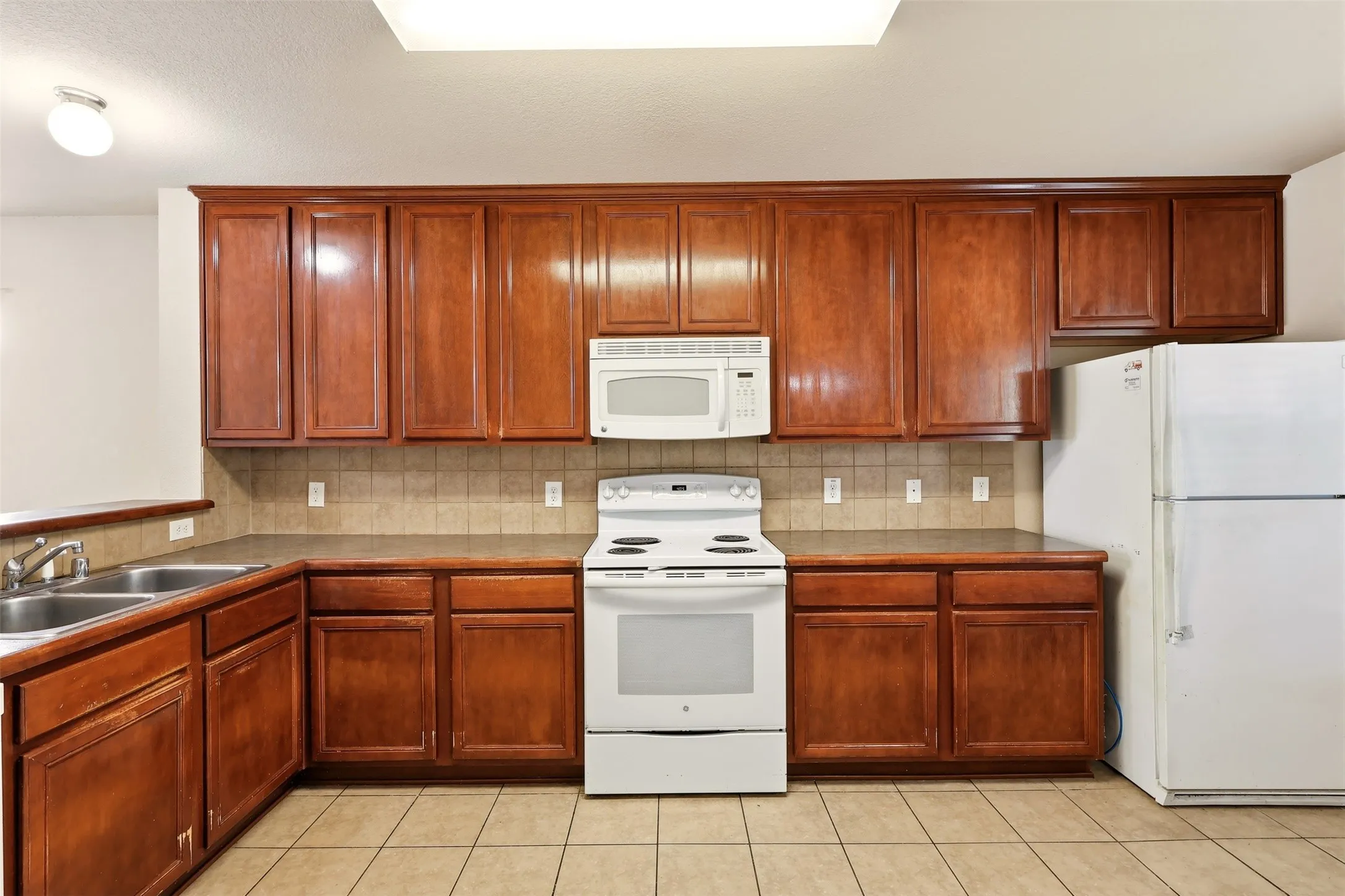 Kitchen featuring wood-finish cabinetry, tile flooring, tile backsplash, laminate countertops, and a double basin stainless steel sink
