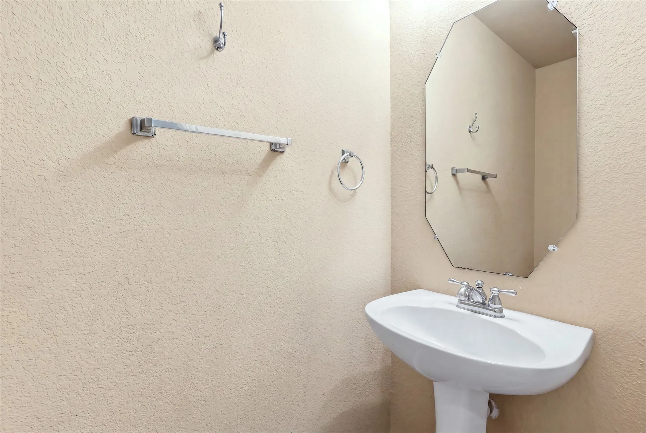 Pedestal sink with dual-handle chrome faucet, octagonal wall mirror, and textured wall finish