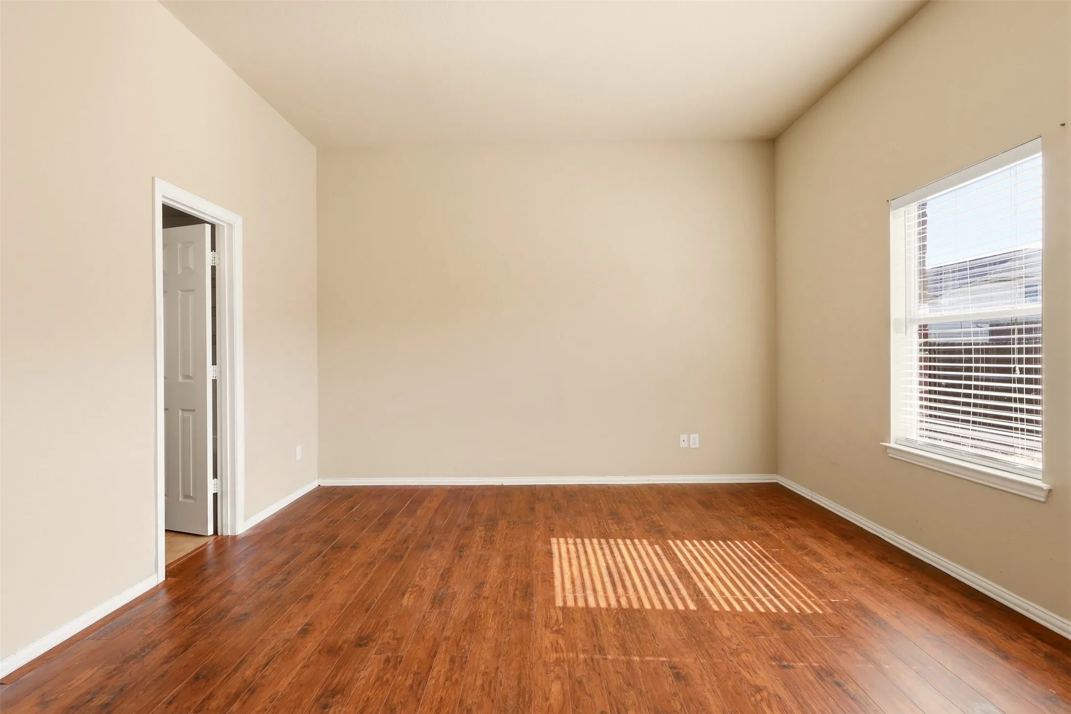 Spacious bedroom with rich wood-finish flooring, abundant natural light from large window with plantation shutters, and neutral wall finishes
