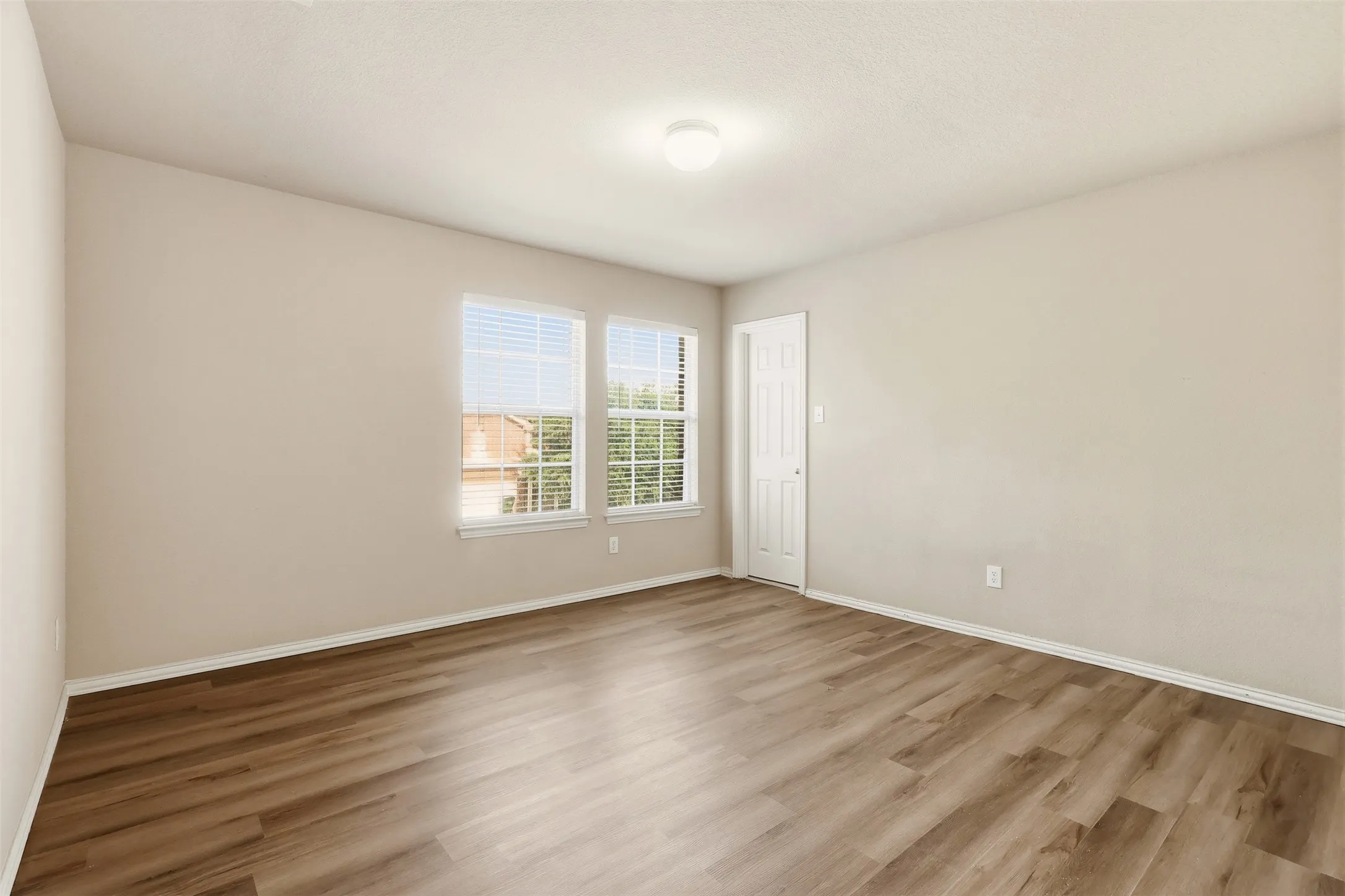 Neutral-toned room featuring two windows with white blinds, wood-finish flooring, and a white paneled door