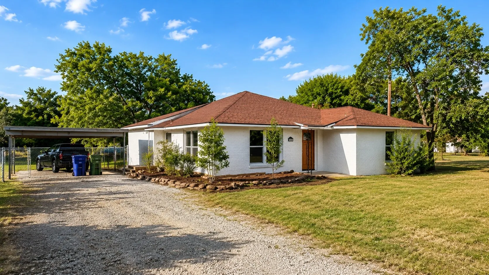 White brick exterior with a red shingle roof, gravel driveway, covered carport, and established landscaping