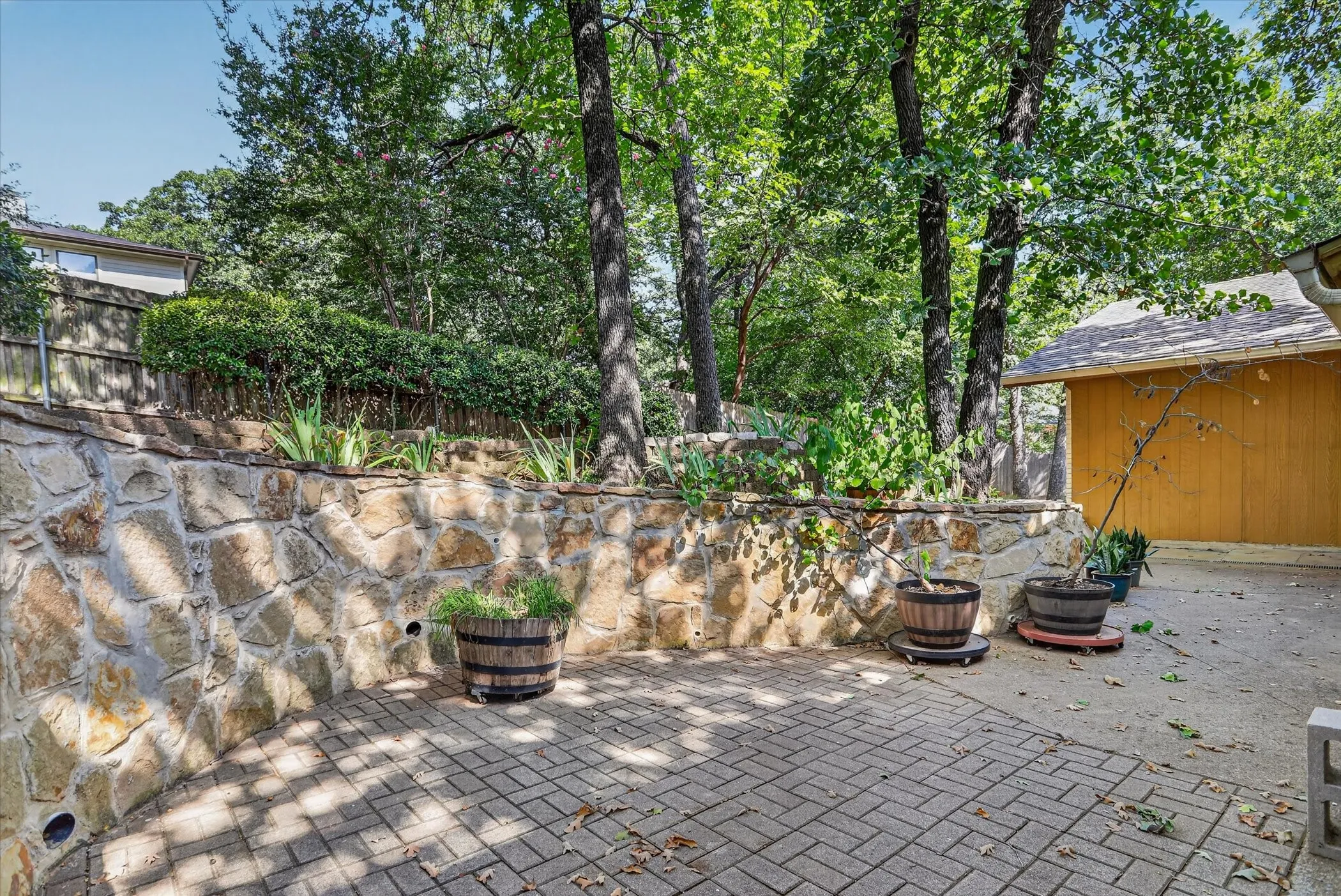 Paver patio with a stone retaining wall, raised planting beds, and mature trees