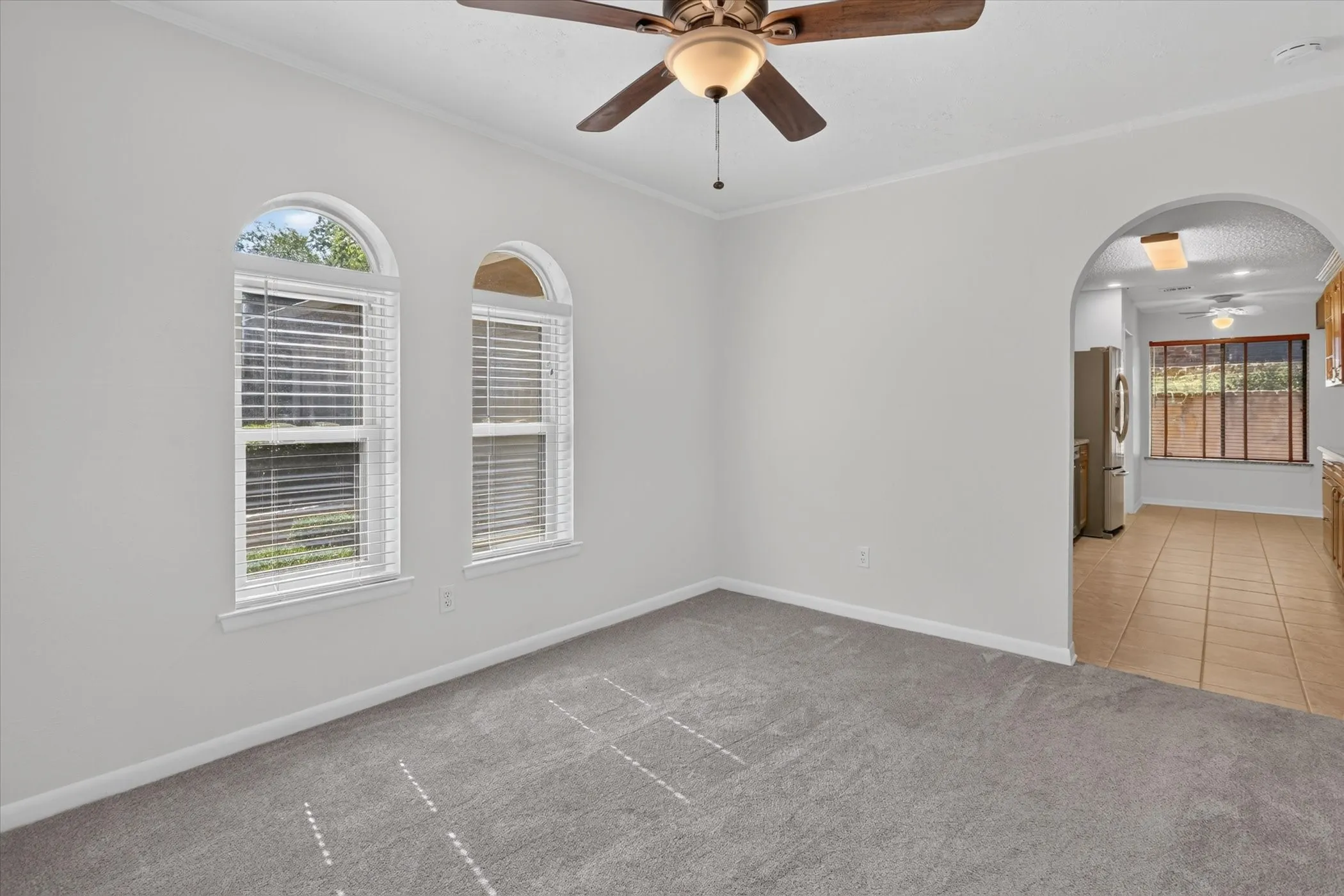 Arched windows with blinds, gray carpeting, ceiling fan with integrated lighting, and an arched doorway leading to a room with tiled flooring