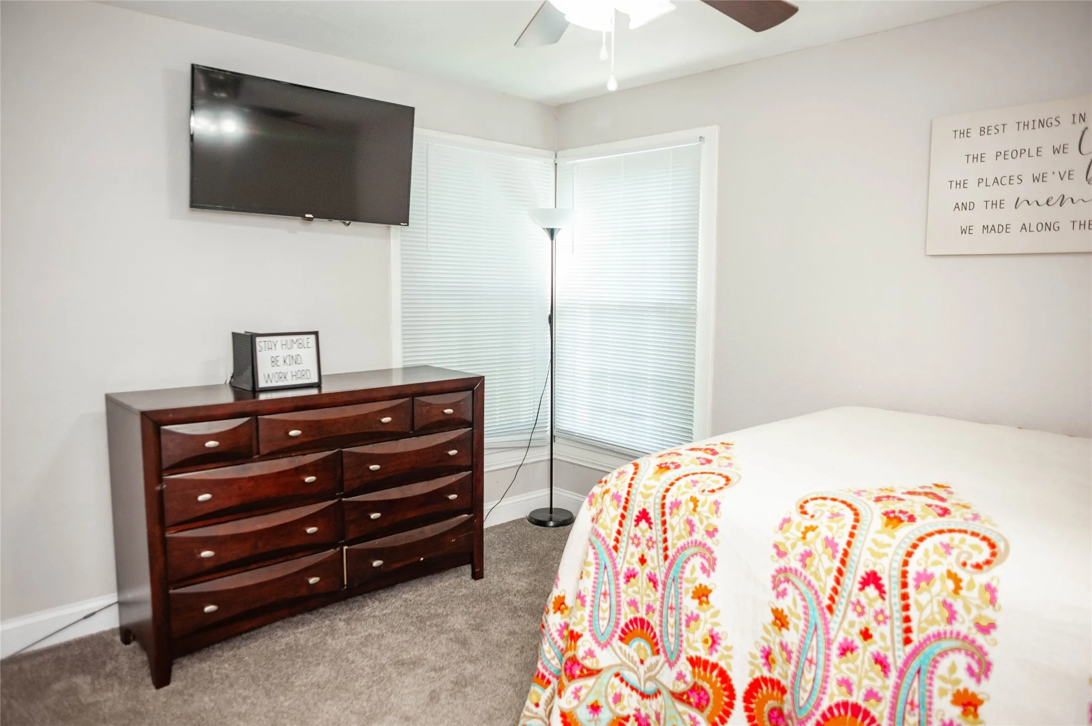 Wall-mounted television fixture, corner windows with blinds, ceiling fan, and carpeted flooring