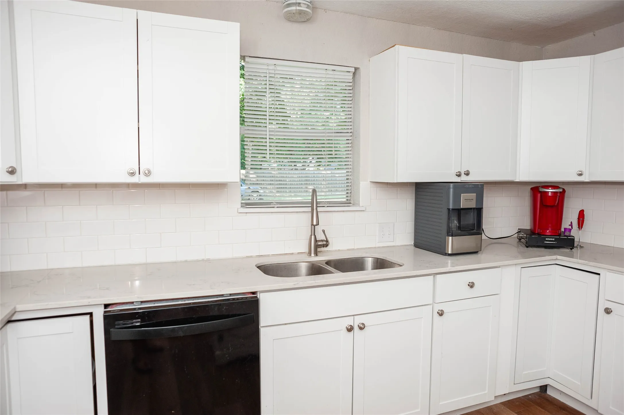 White shaker cabinetry, light-toned countertops, white subway tile backsplash, stainless steel double basin sink, and a stainless steel faucet