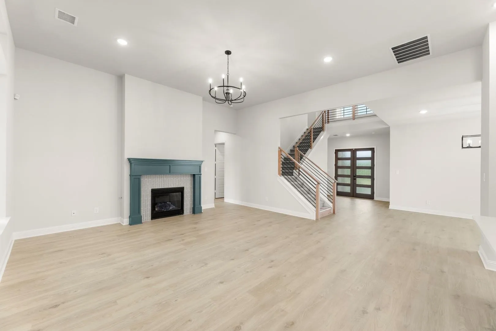 Spacious living area featuring light wood-finish flooring, a built-in fireplace with a teal mantel and tiled surround, and a contemporary chandelier
