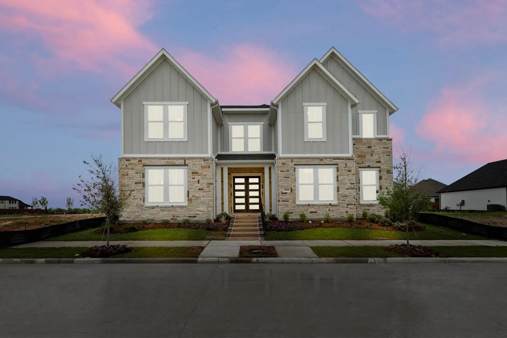 Two-story home with light grey board and batten siding, stone facade accents, a multi-pane front door, white window frames, and a concrete pathway leading to the entrance