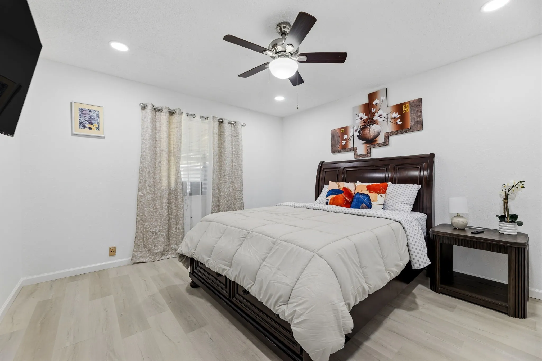 Bedroom featuring light wood-finish flooring, a contemporary ceiling fan with integrated lighting, recessed ceiling lights, a window with patterned drapes, and a dark wood bed frame with a prominent headboard