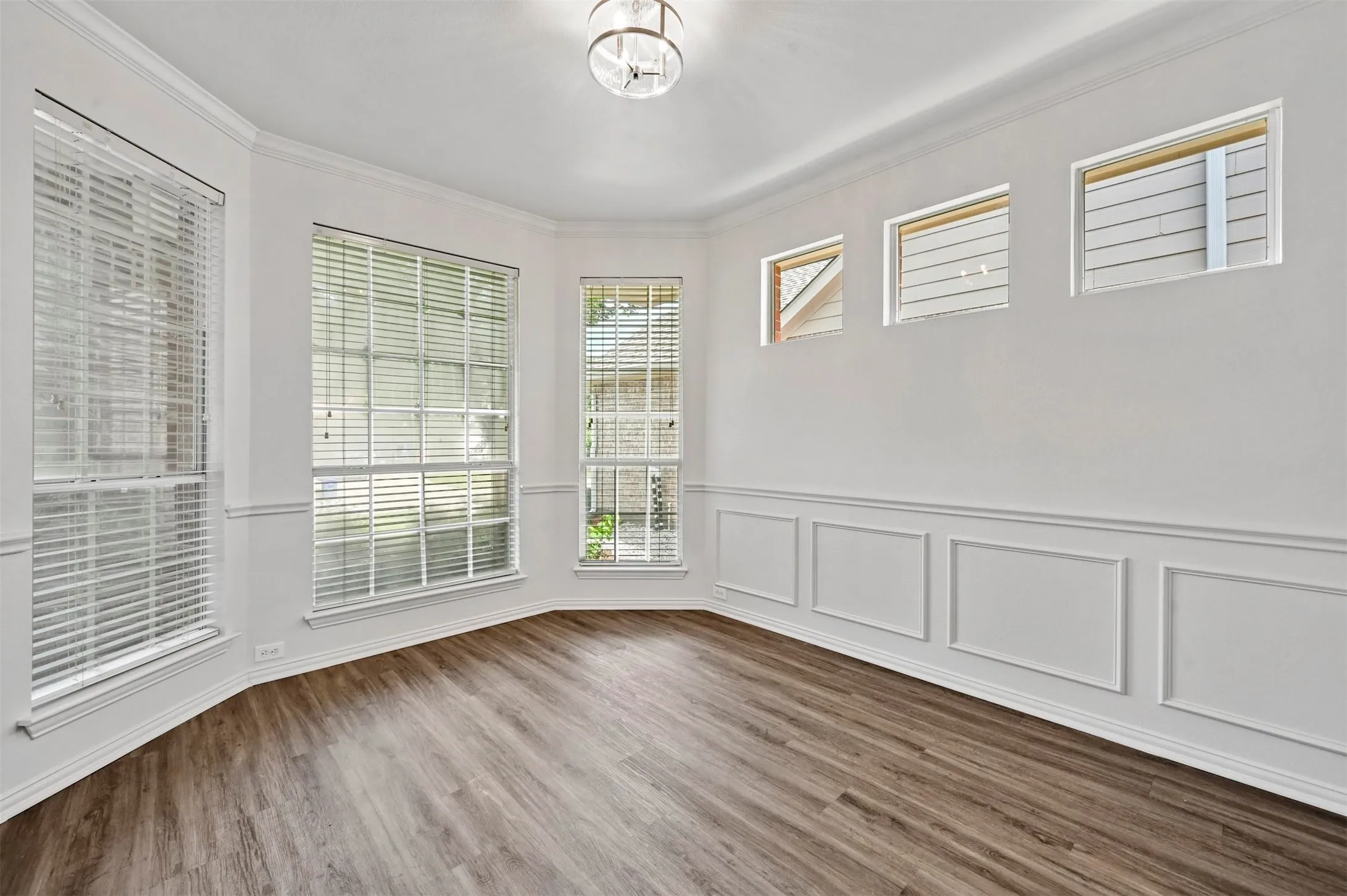 Spacious room featuring wood-finish flooring, wainscoting, crown molding, and multiple windows with blinds
