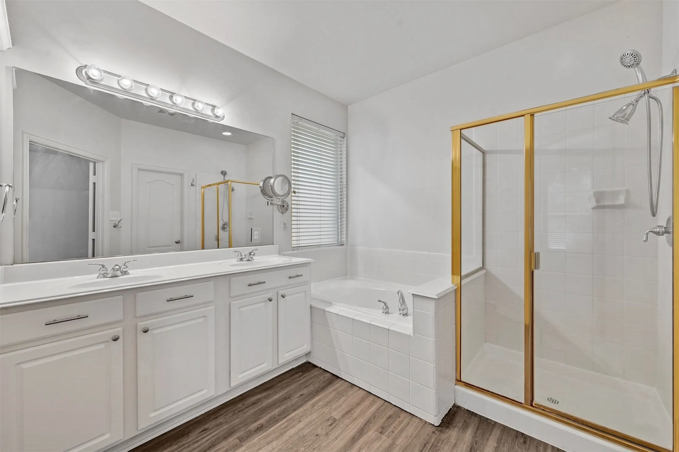Bathroom featuring a double vanity with white cabinetry, wood-finish flooring, a built-in soaking tub, and a separate shower with a gold-framed glass enclosure