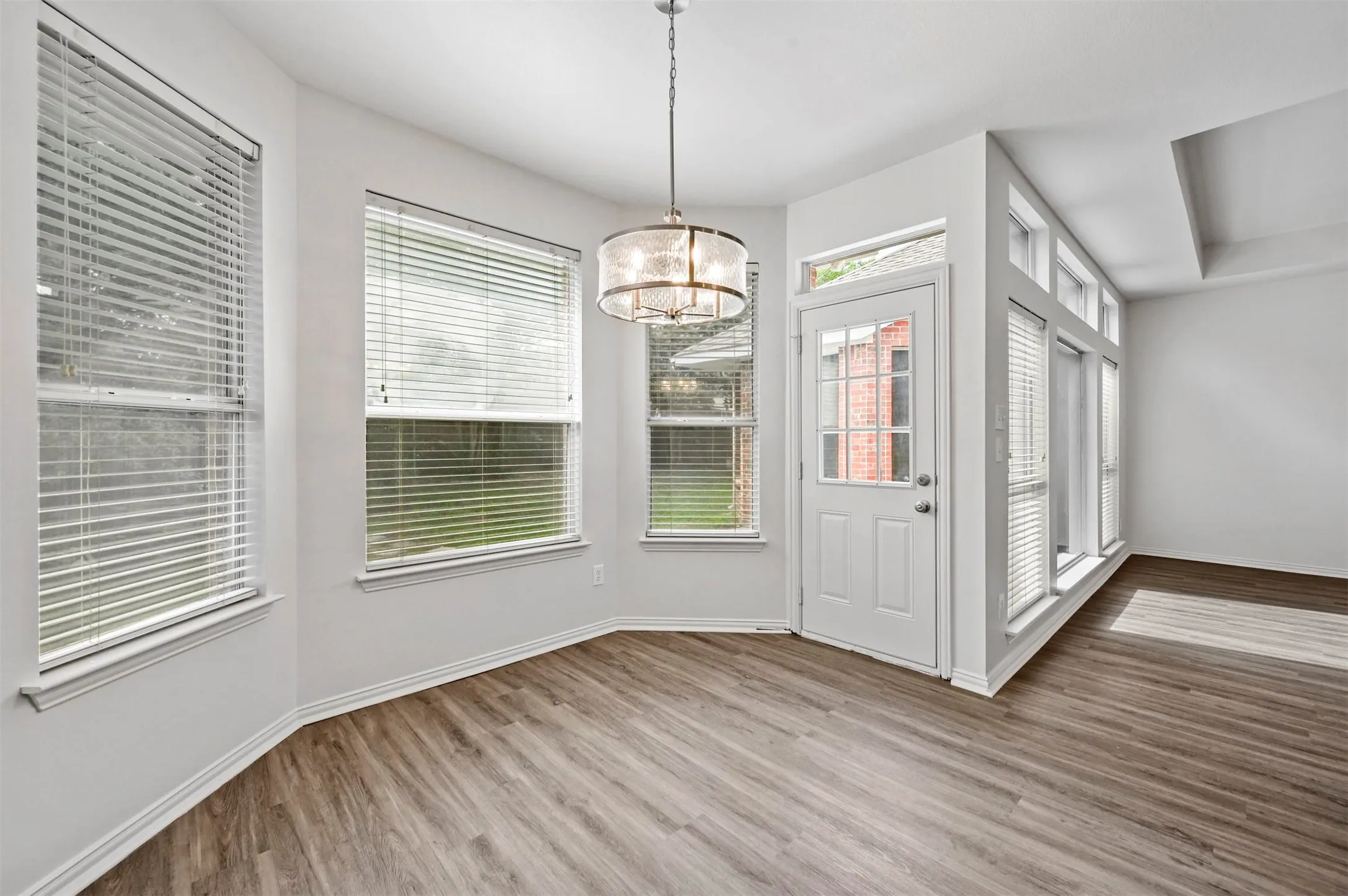 Bay window dining area featuring wood-finish flooring, a modern drum chandelier, multiple windows with blinds, a white paneled door with glass inserts, and white baseboards