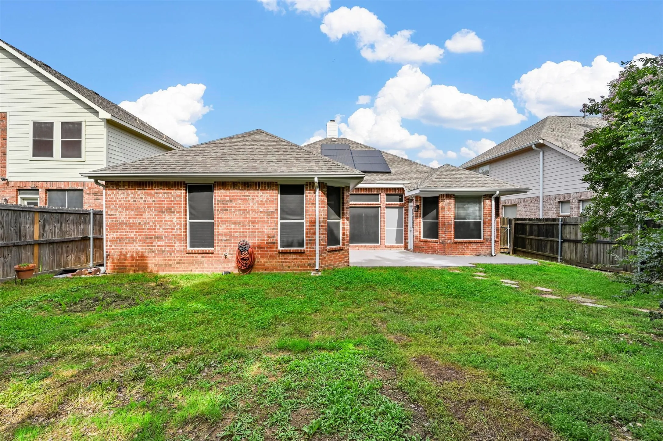 Spacious backyard featuring a concrete patio, brick exterior, multiple windows, solar panels, and wood fencing