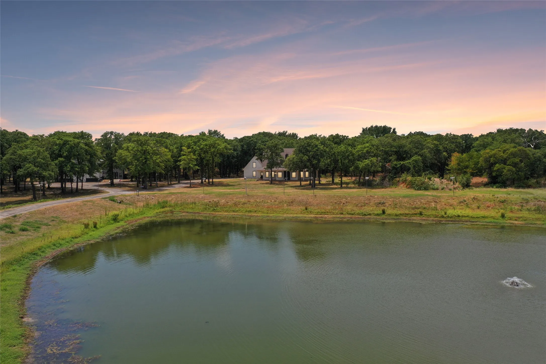 Expansive property featuring a tranquil pond with a fountain, surrounded by mature trees, and a gravel drive leading to a two-story residence