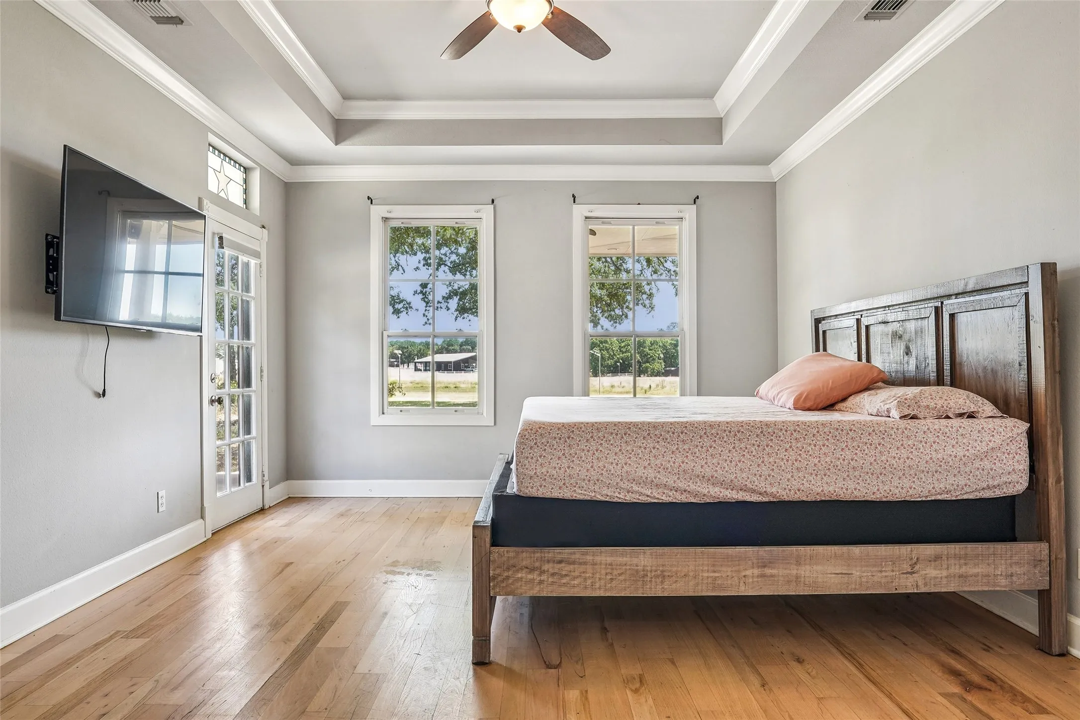 Tray ceiling with crown molding, natural wood-finish flooring, two large windows, a glass-paneled door, and a ceiling fan with integrated lighting
