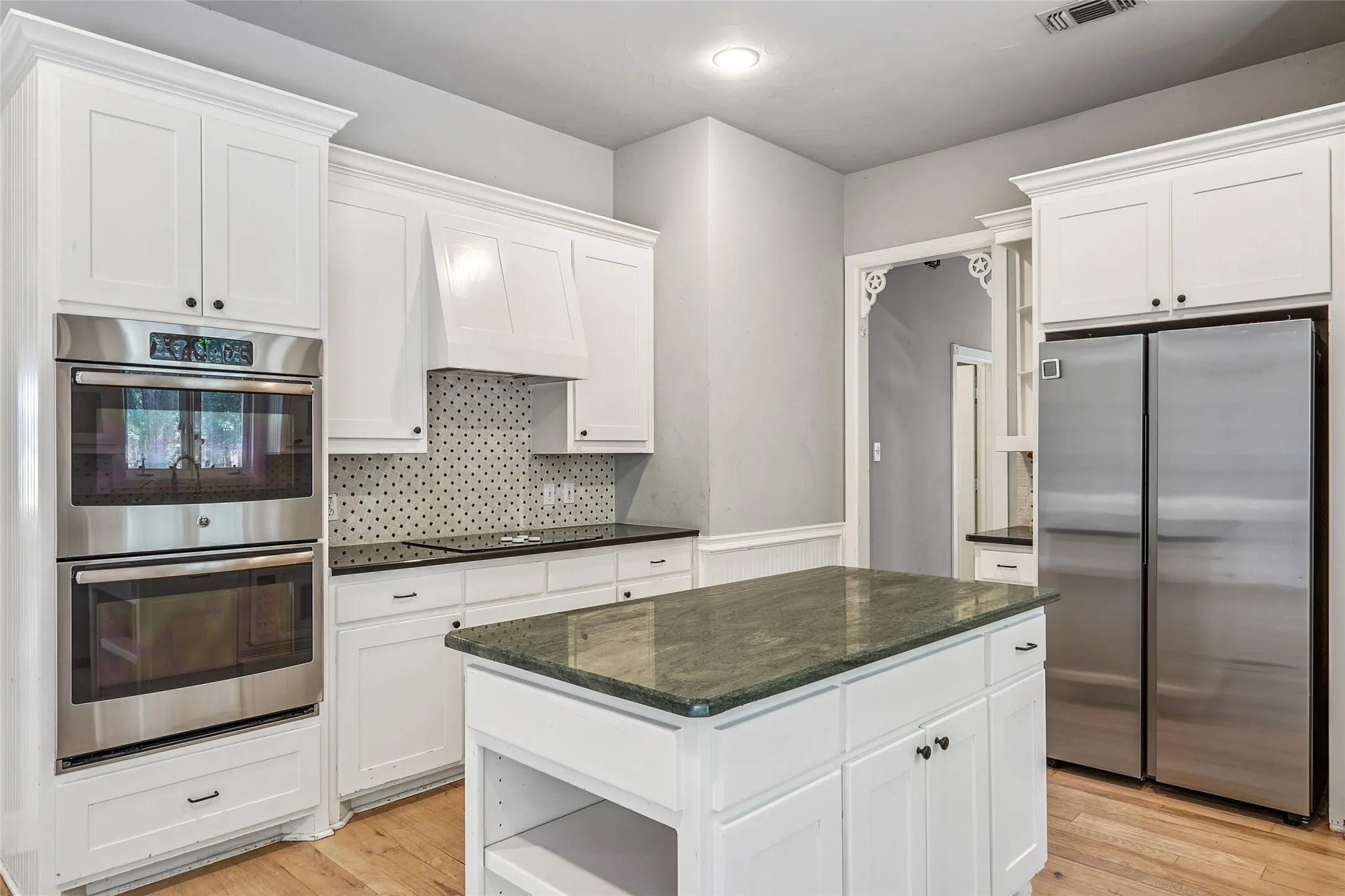 Contemporary kitchen featuring white cabinetry, stainless steel double wall ovens, a dark stone countertop, a tiled backsplash, and a central island with a green stone surface