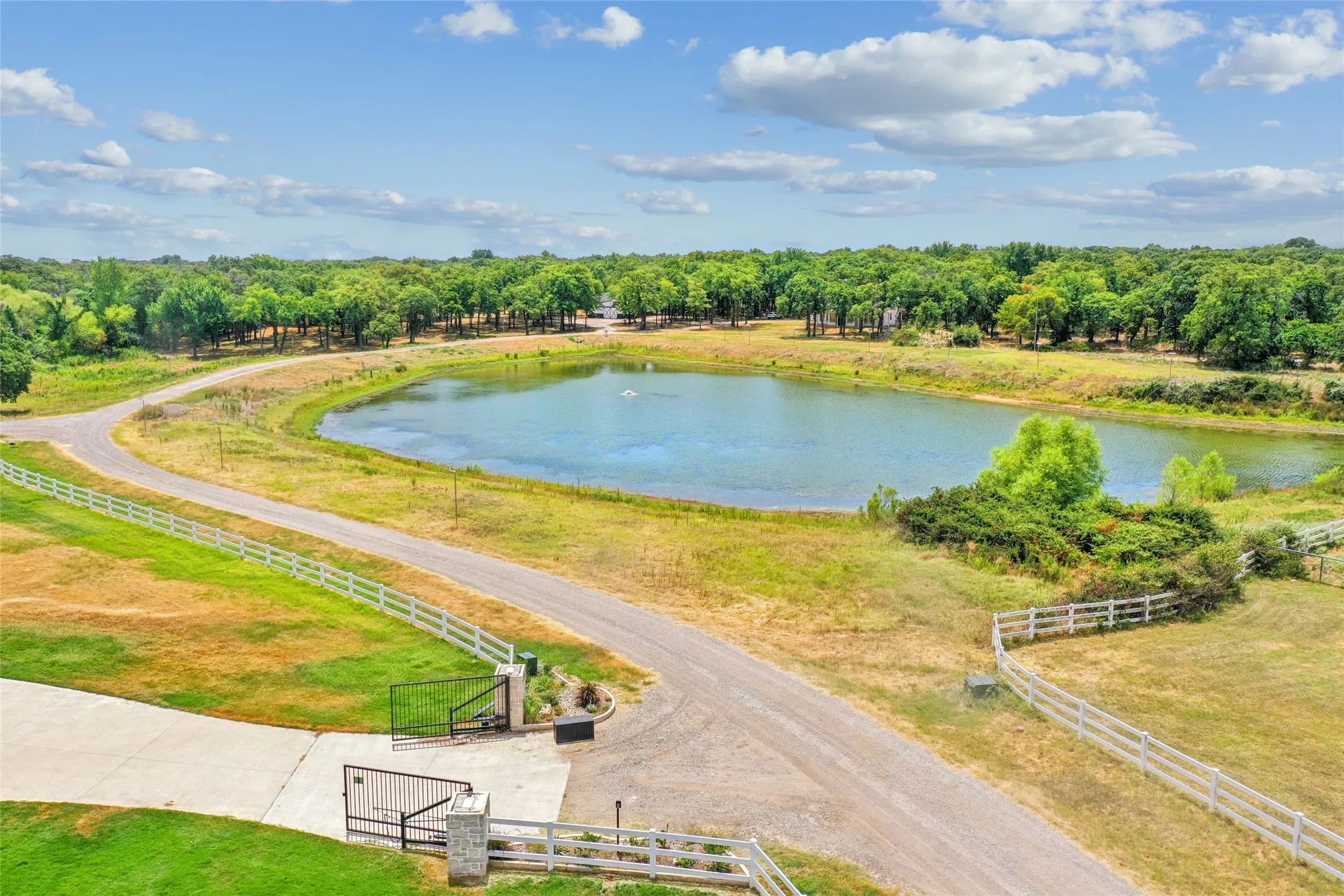 Private gated entrance opening to expansive grounds featuring a large pond with a water feature, perimeter fencing, and mature trees creating a natural backdrop