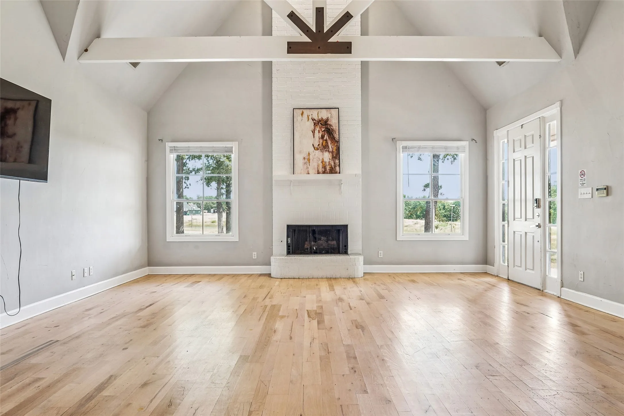 Spacious living area featuring a vaulted ceiling with exposed beams, light wood-finish flooring, and a white-painted brick fireplace