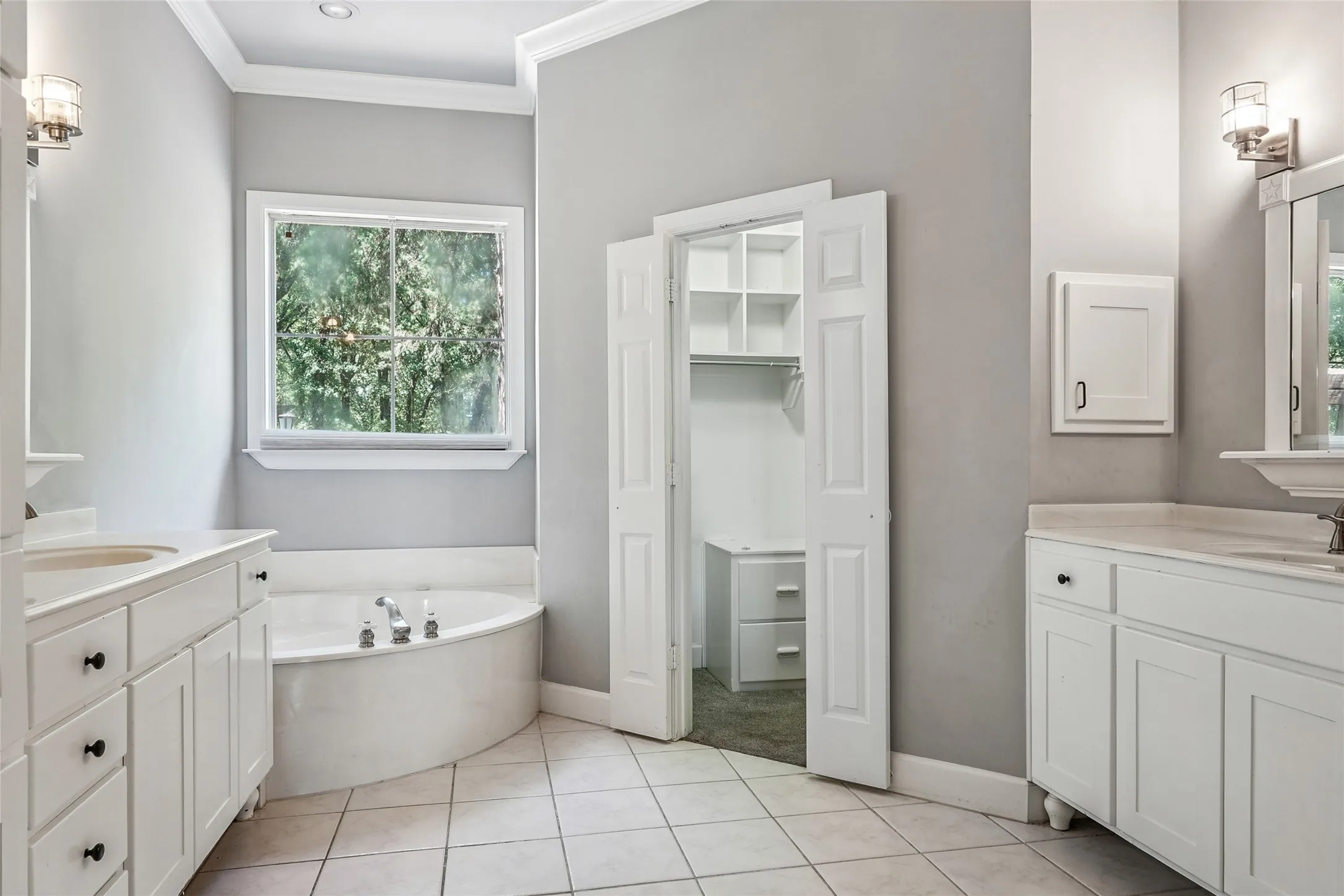 Bathroom featuring a built-in corner soaking tub, dual white vanity cabinets with integrated sinks, and neutral tile flooring