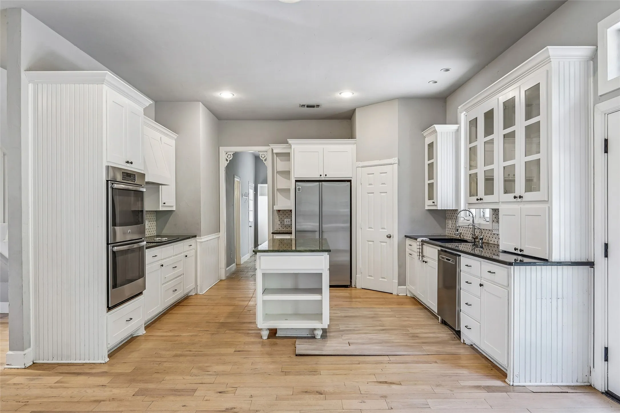 Spacious kitchen with white cabinetry, black countertops, stainless steel appliances including double ovens and a refrigerator, and wood-finish flooring